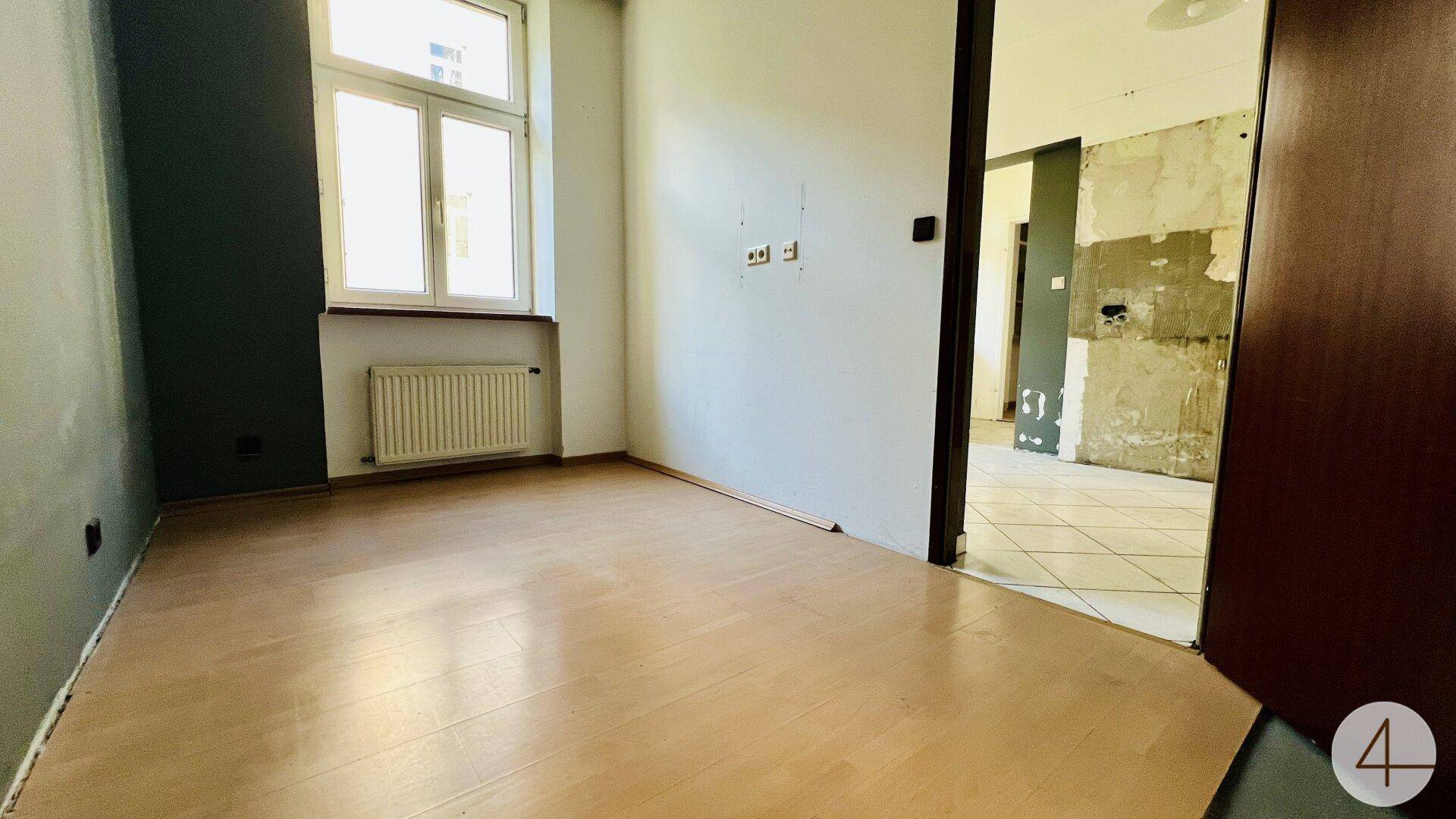 Leeres Zimmer mit Holzfußboden und Fenster, angrenzend an einen Renovierungsbereich.