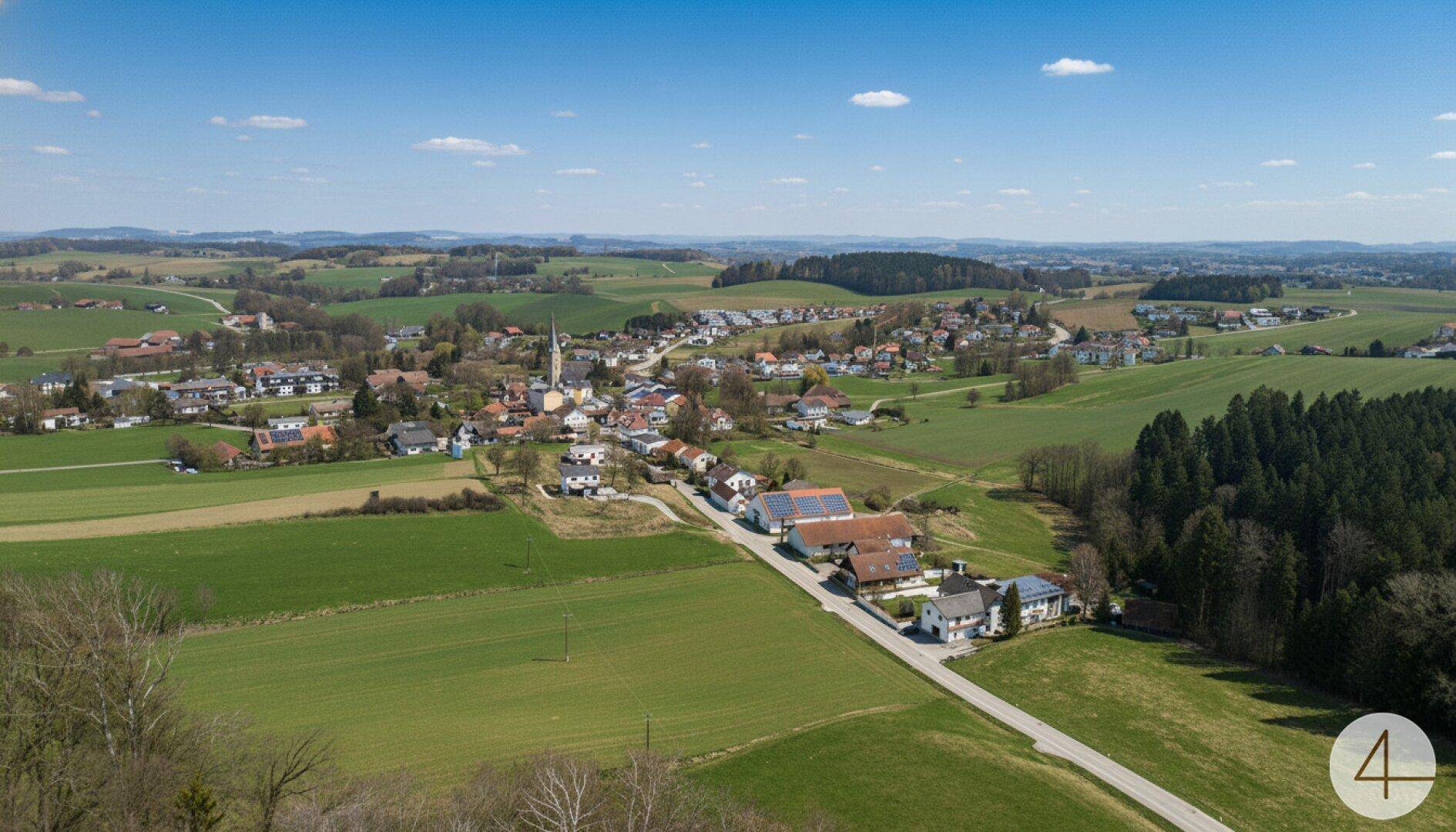 Panoramablick aus der Luft auf ein Dorf mit Kirche, umgeben von grünen Feldern und Hügeln.