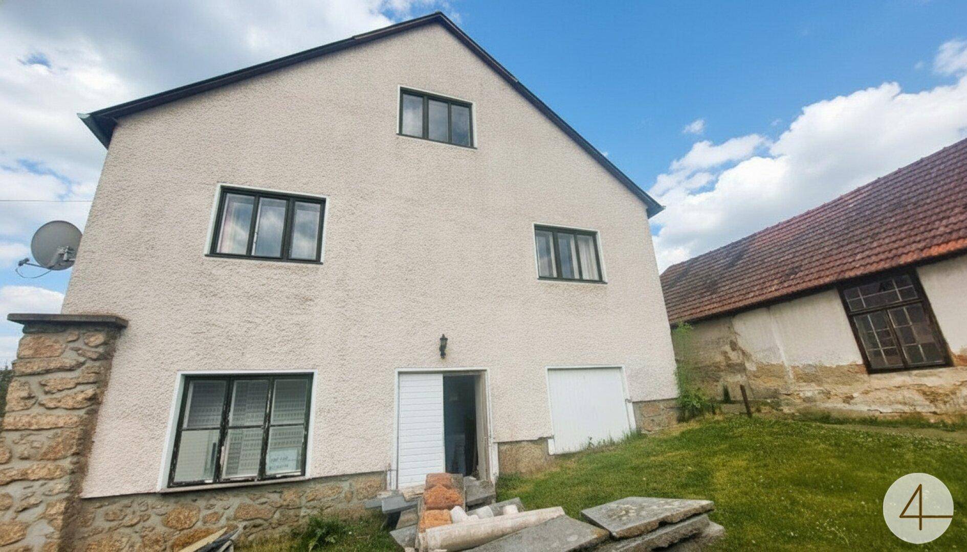 Seitenansicht eines Hauses mit Putzfassade und angrenzendem Nebengebäude unter blauem Himmel.