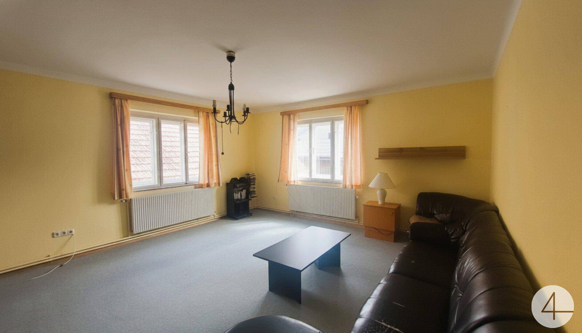 Wohnzimmer mit Teppichboden, Ledersofa, Couchtisch und zwei Fenstern mit Heizkörpern.