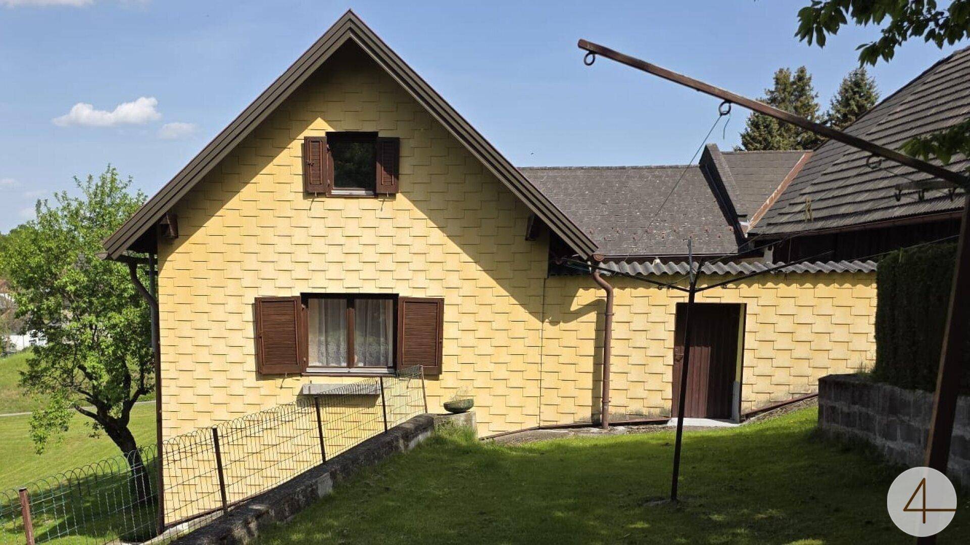 Gelbes Haus mit braunen Fensterläden und einem grünen Gartenbereich mit Zaun.