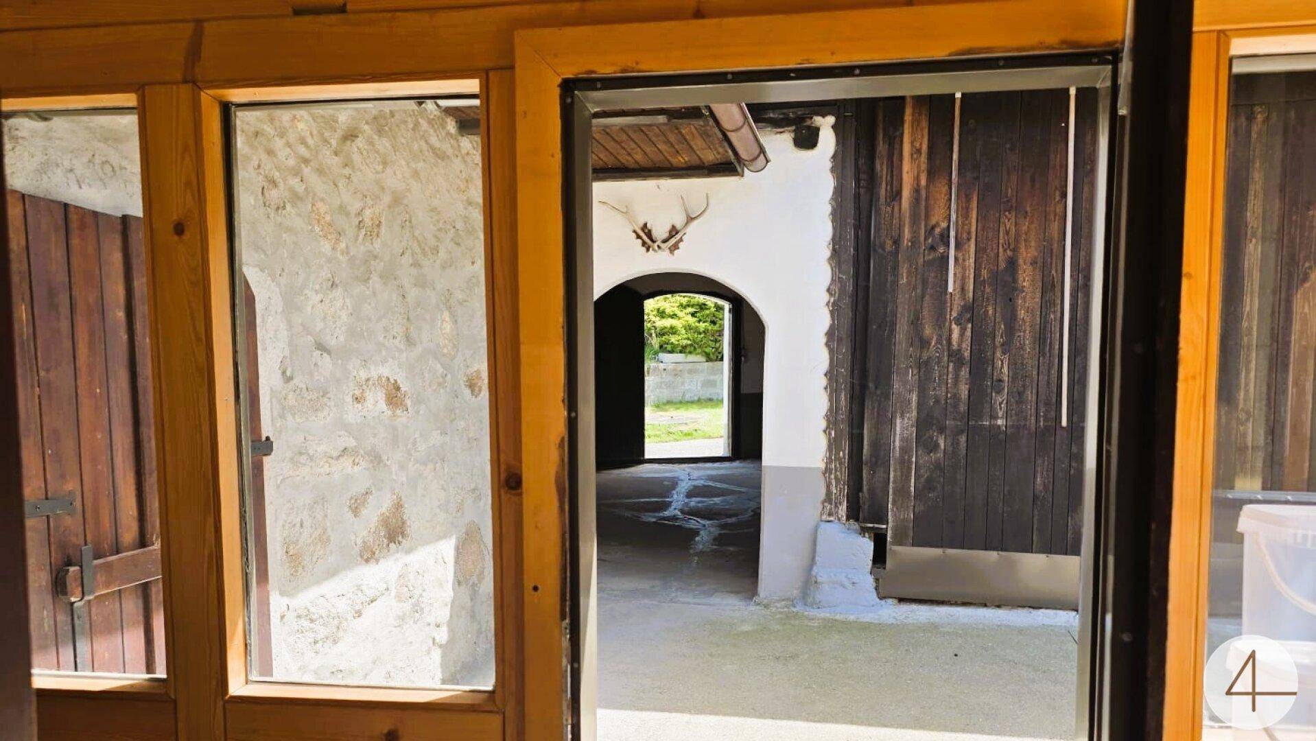 Blick aus einem Innenraum in einen rustikalen Innenhof mit Steinmauern und Holzelementen.