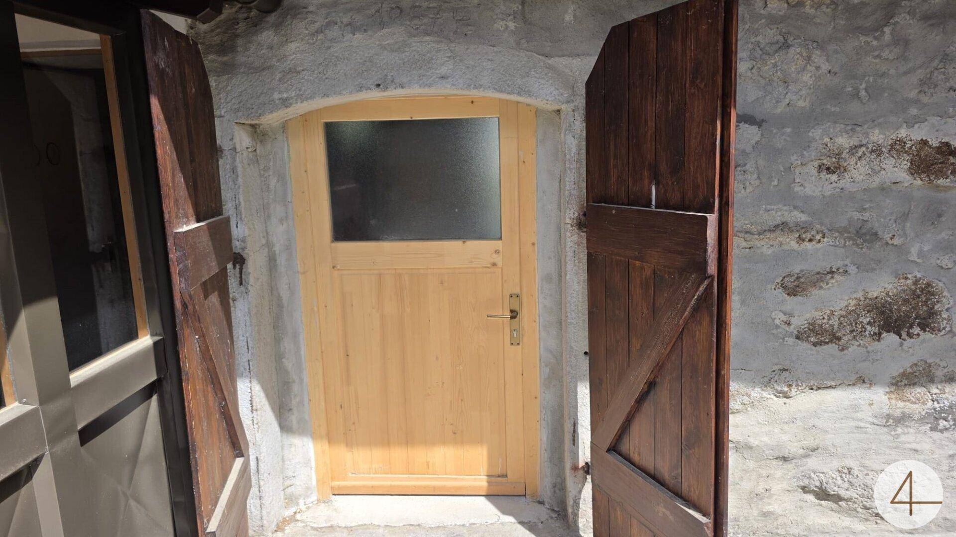Rustikale Holztür mit Glasausschnitt, umgeben von Steinmauern und hölzernen Fensterläden.