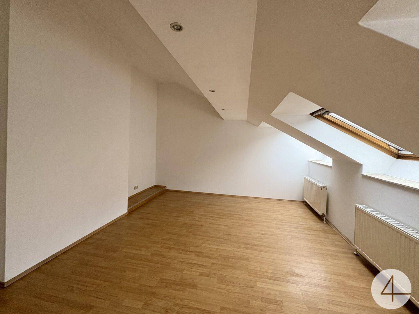 Heller Raum im Dachgeschoss mit Holzfußboden, schrägen Wänden und einem Dachfenster.