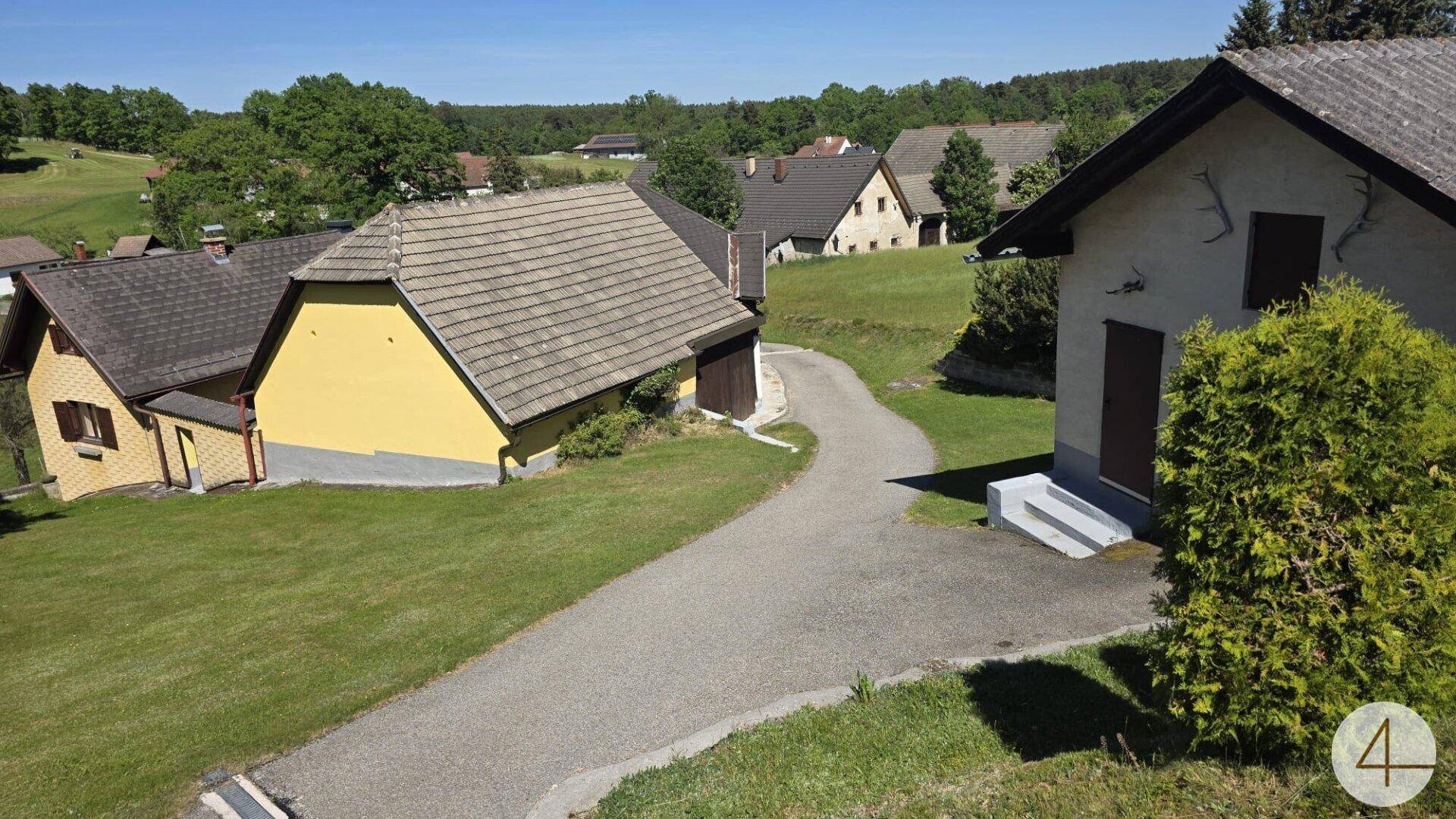 Ländliche Ansicht mit einer Zufahrt, mehreren Häusern und üppiger Vegetation an einem sonnigen Tag.