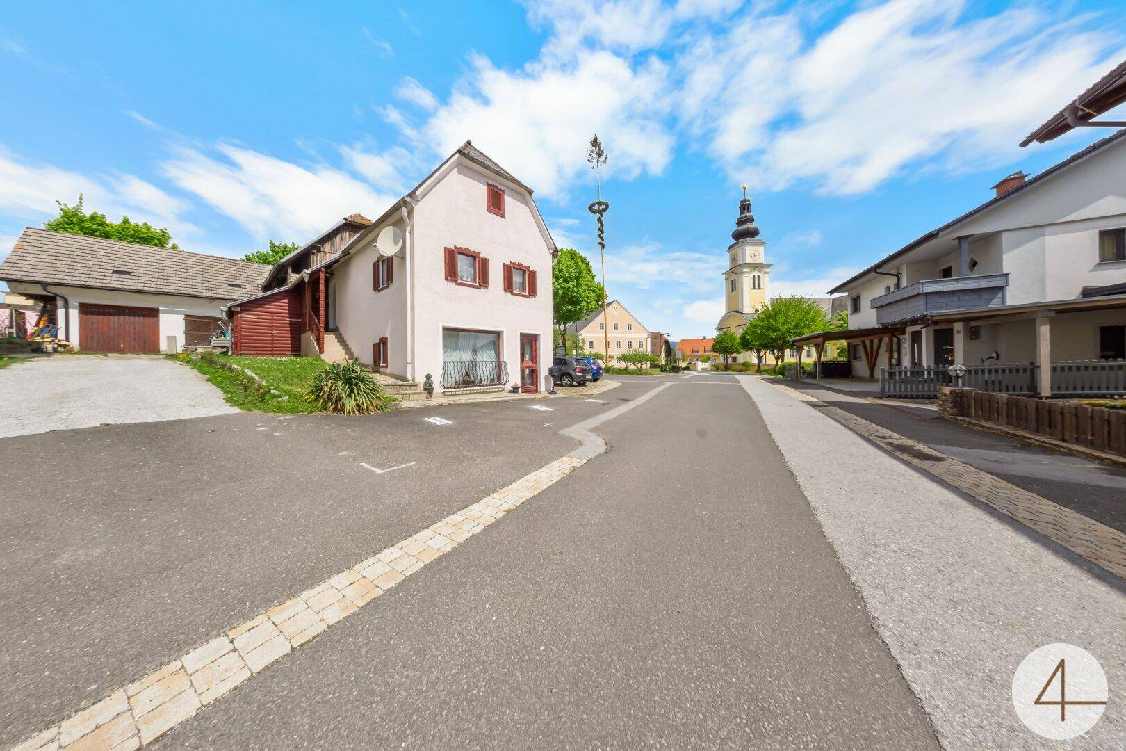 Traditionelles Gebäude an einer Dorfstraße mit Kirchturm im Hintergrund unter blauem Himmel.