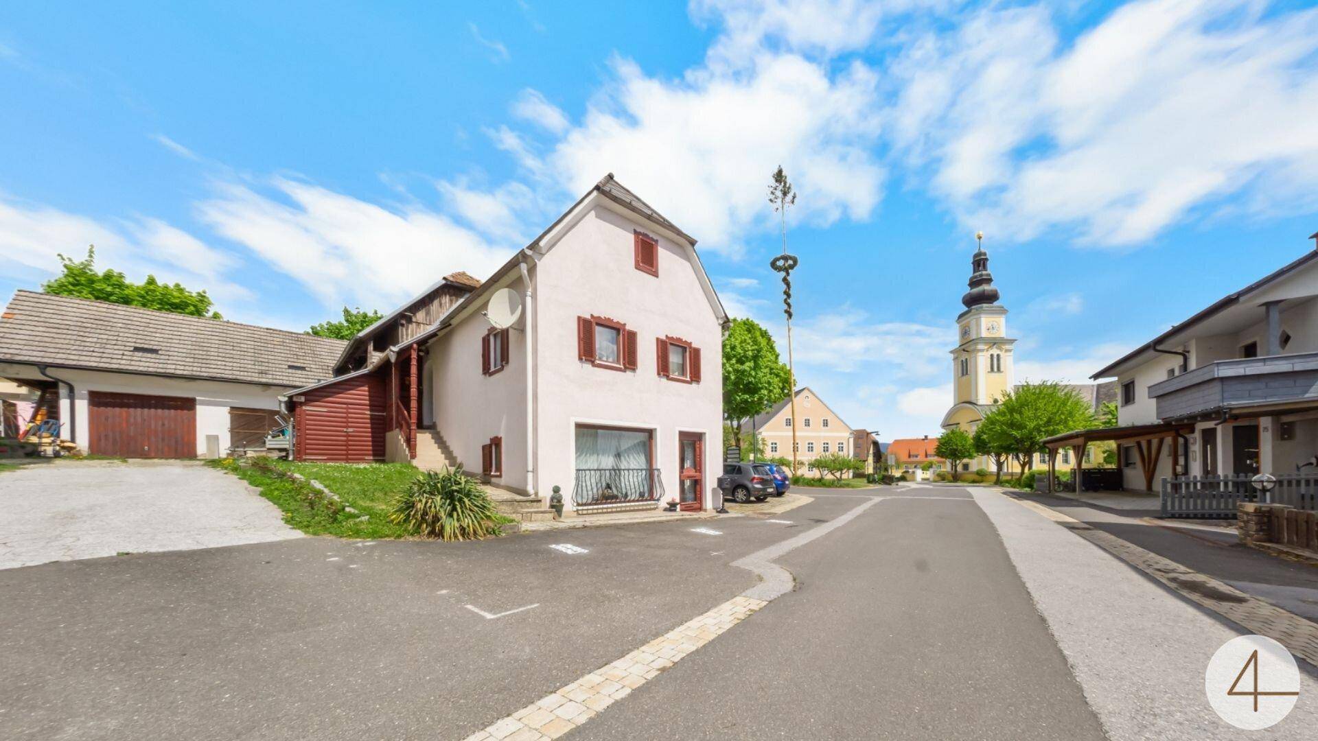 Traditionelles Gebäude an einer Dorfstraße mit Kirchturm im Hintergrund unter blauem Himmel.