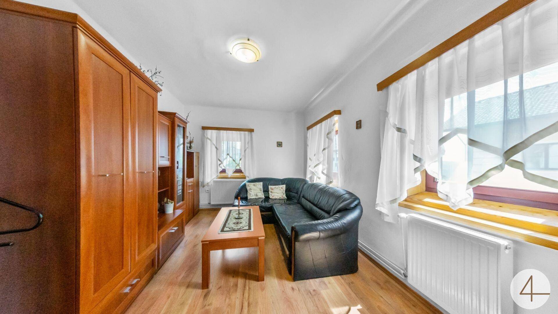 Wohnzimmer mit schwarzem Ledersofa, Holzmöbeln und hellen Vorhängen an den Fenstern.