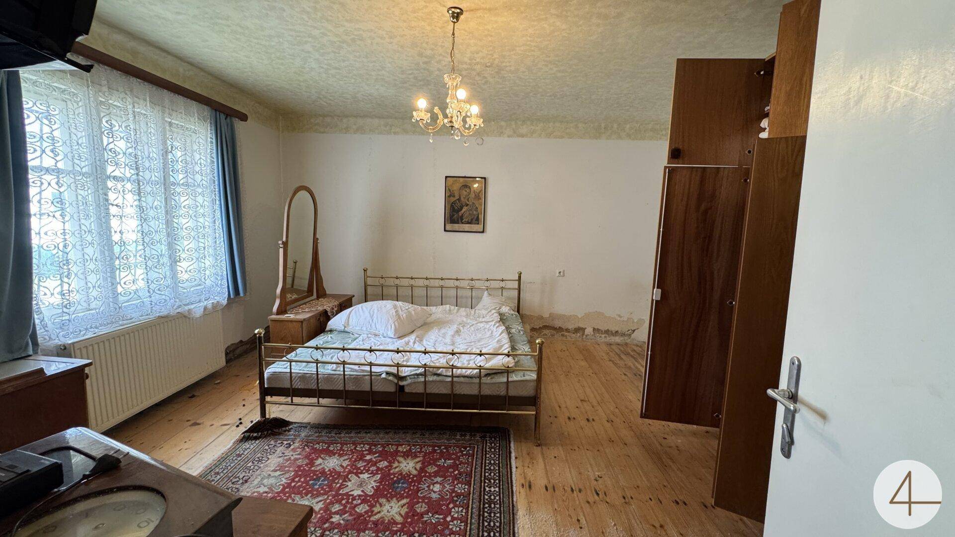 Schlafzimmer mit Metallbett, großem Fenster, Kronleuchter und Holzdielenboden.