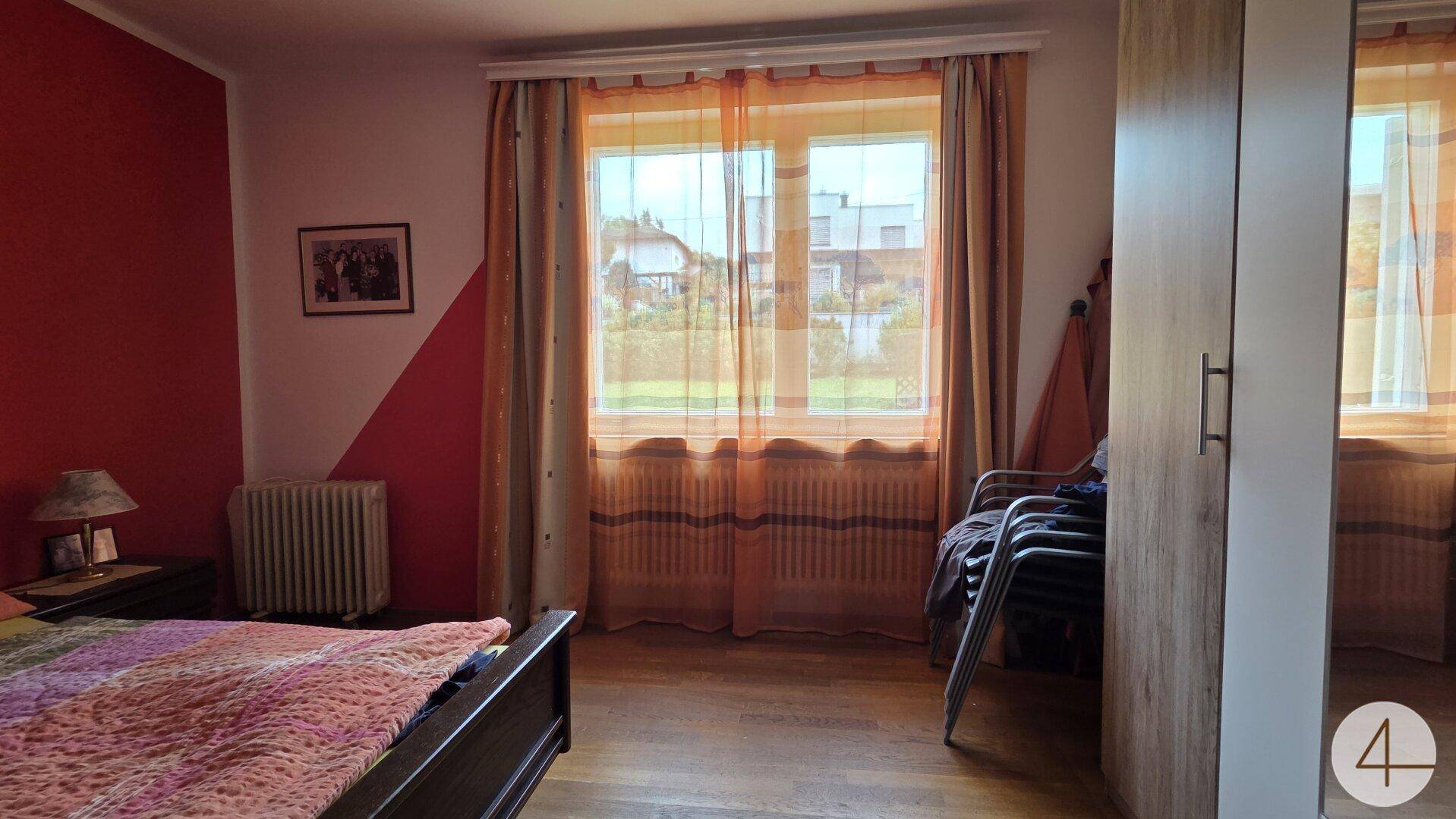 Schlafzimmer mit Holzboden, großem Fenster und farbigen Wänden in Rot- und Beigetönen.