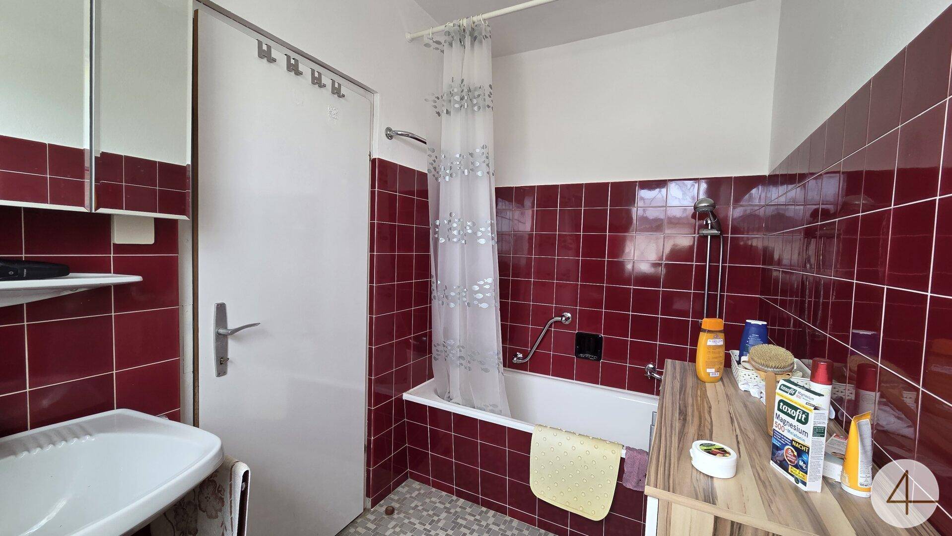 Badezimmer mit roten Fliesen, Badewanne mit Duschvorhang und einem Waschbecken im 80er-Jahre-Stil.