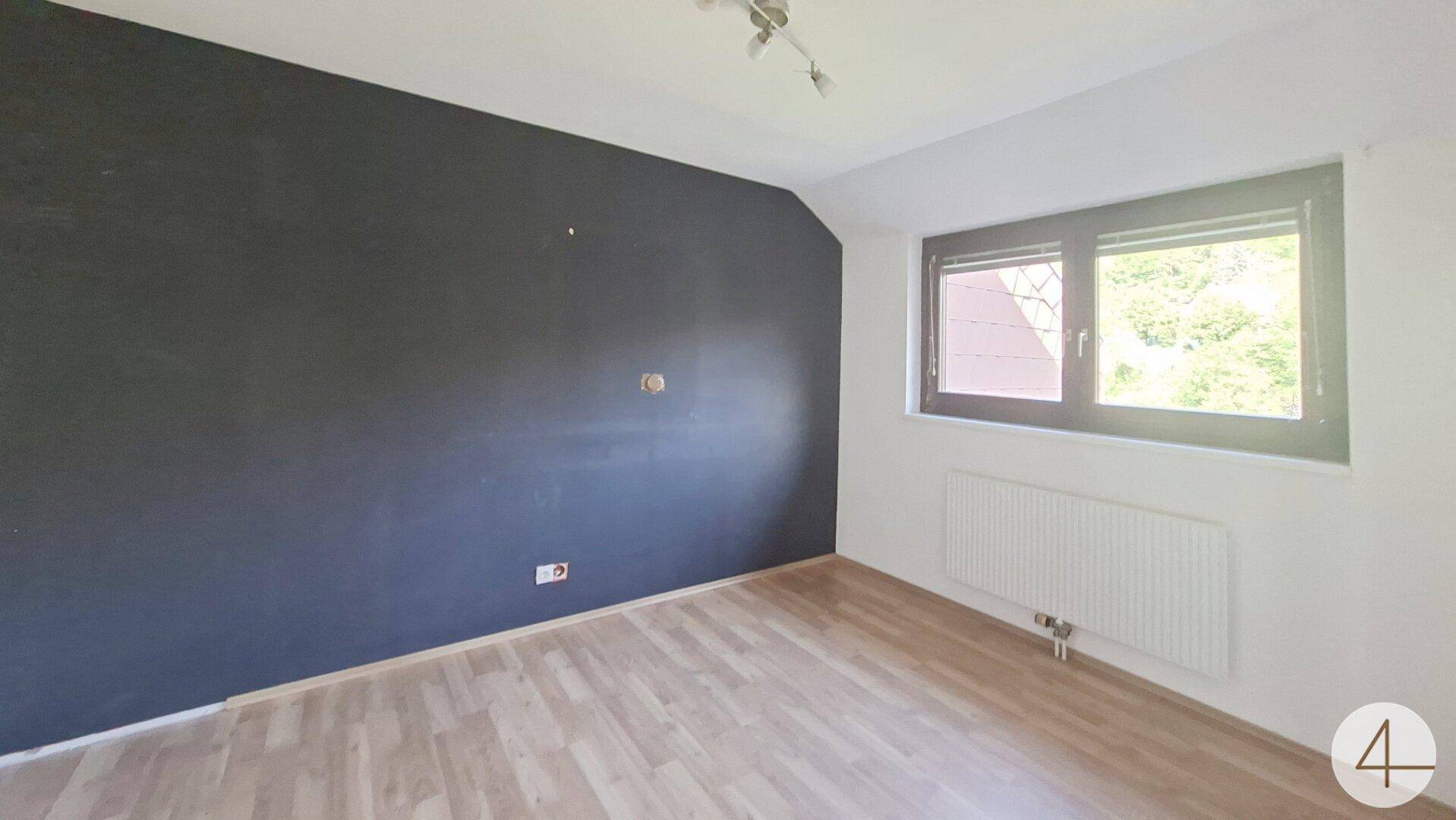 Schlafzimmer mit dunkler Akzentwand, Laminatboden und Fenster mit Blick ins Grüne.