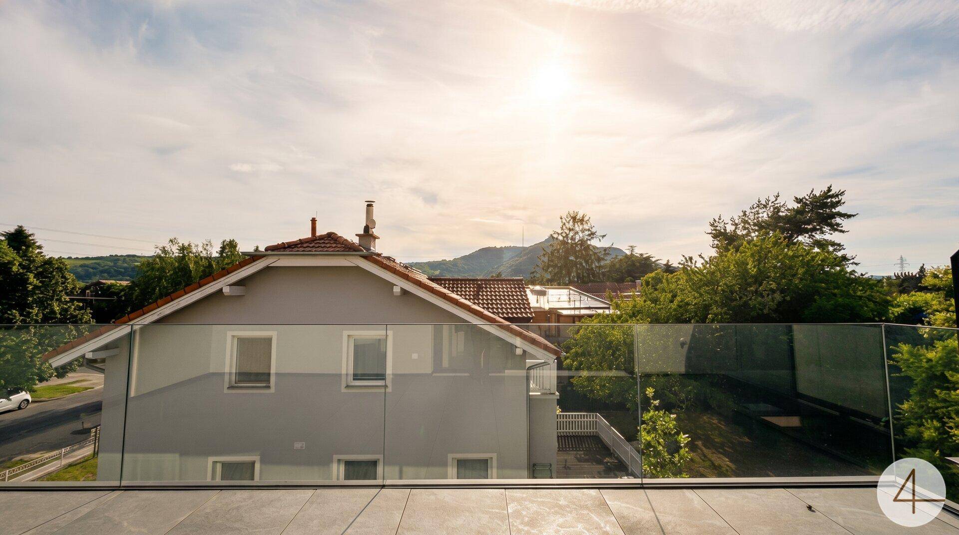 Sonniger Ausblick von einer Terrasse mit Glasgeländer auf Dächer, Bäume und Berge.