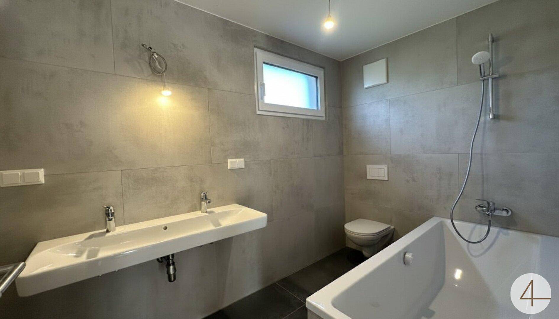Modernes Badezimmer mit grauen Fliesen, Doppelwaschbecken, Badewanne und Fenster.