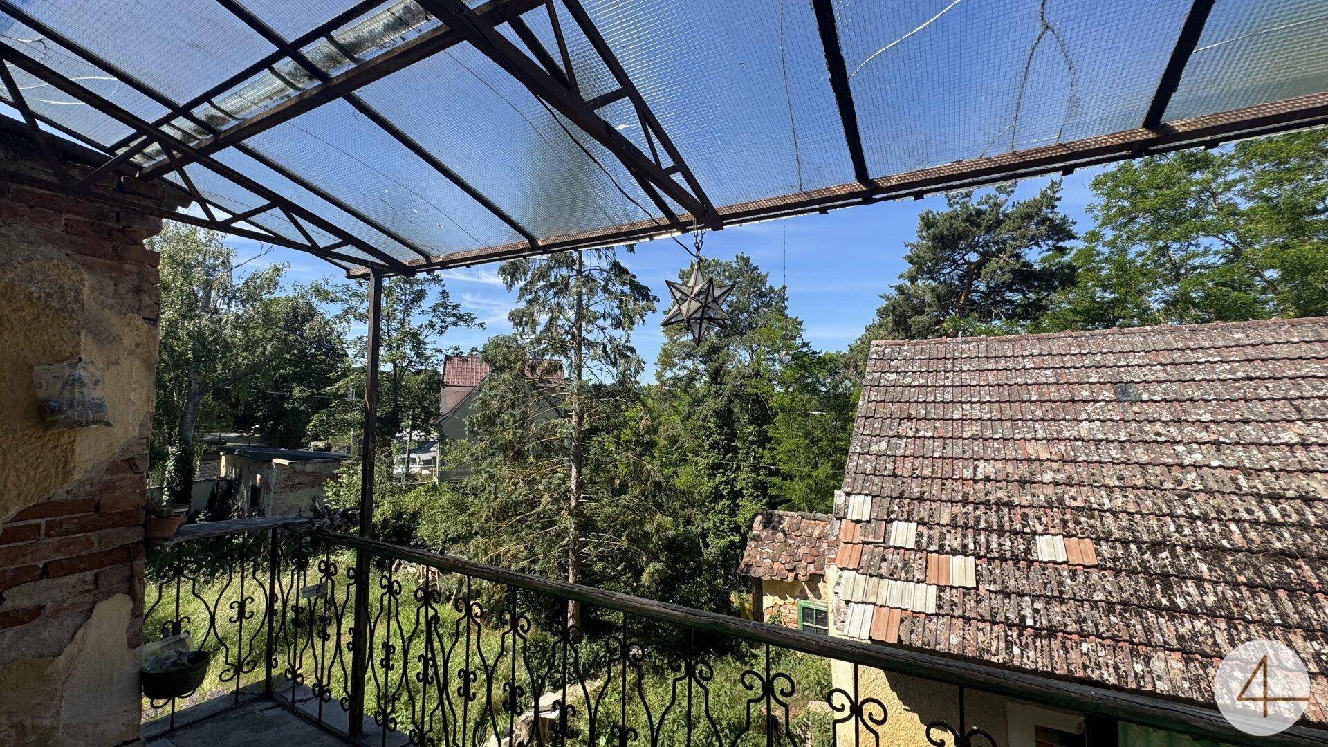 Überdachter Balkon mit schmiedeeisernem Geländer und Blick auf ein Ziegeldach sowie grüne Bäume.