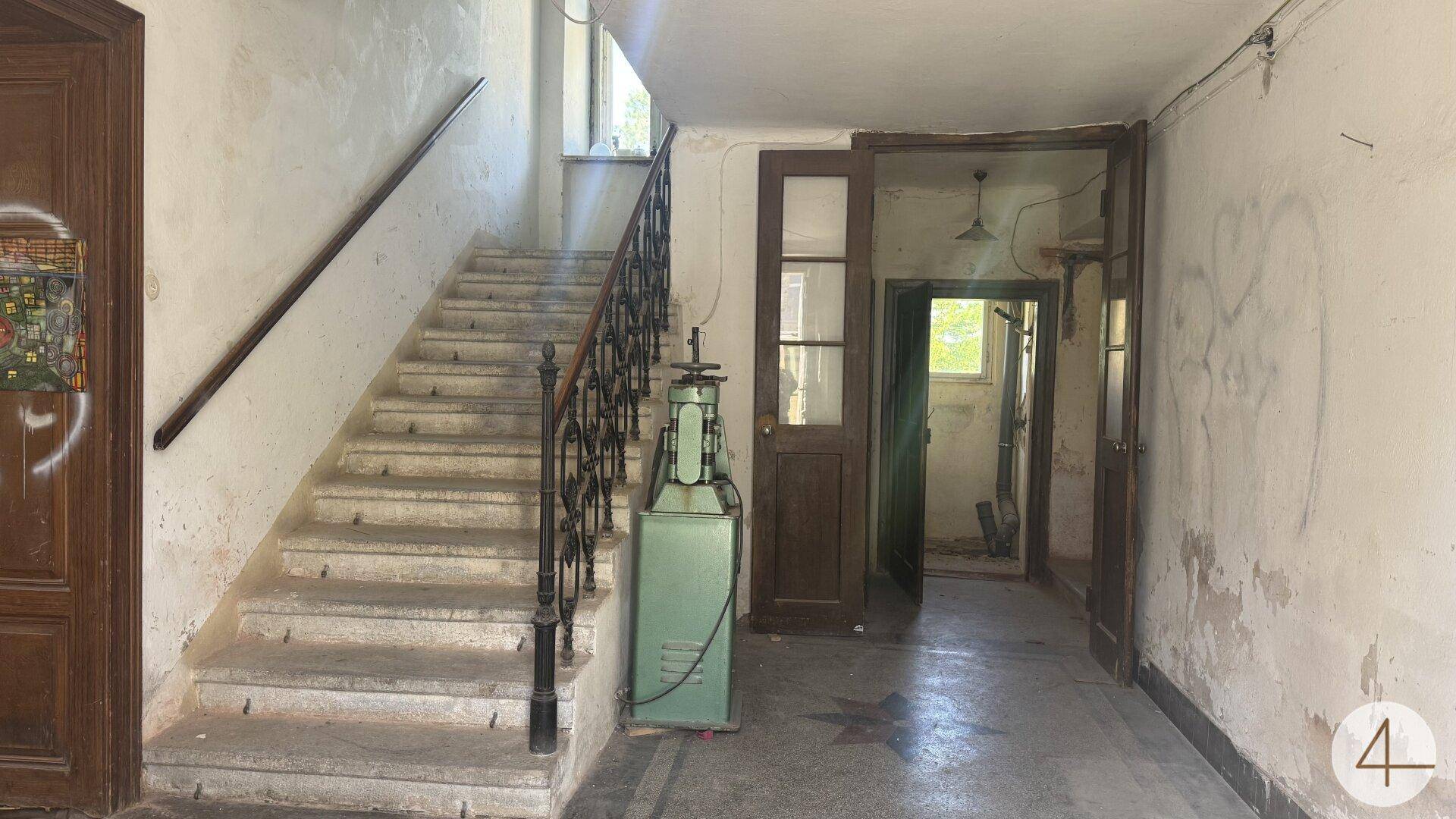 Alter Flur mit abgenutzter Steintreppe und schmiedeeisernem Geländer, der Renovierungsbedarf aufweist.