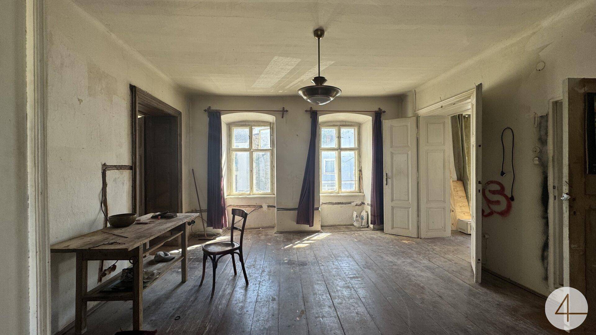 Großes, helles Zimmer mit Holzdielenboden, zwei Fenstern und einfacher Einrichtung, das Renovierung benötigt.