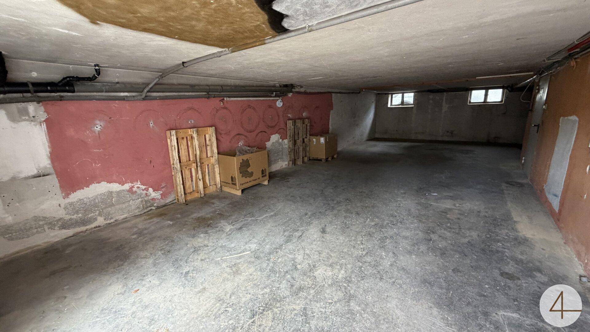 Kellerraum mit Betonboden und rot gestrichenen Wänden, kleinen Fenstern nahe der Decke.