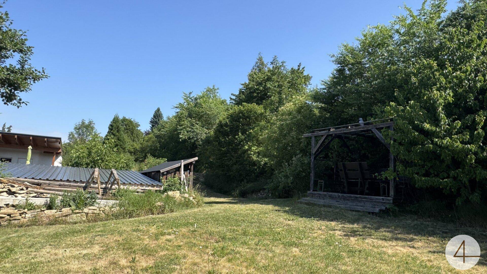 Weitläufiger Gartenbereich mit Rasenfläche, Bäumen und einem Holzpavillon im Hintergrund unter blauem Himmel.