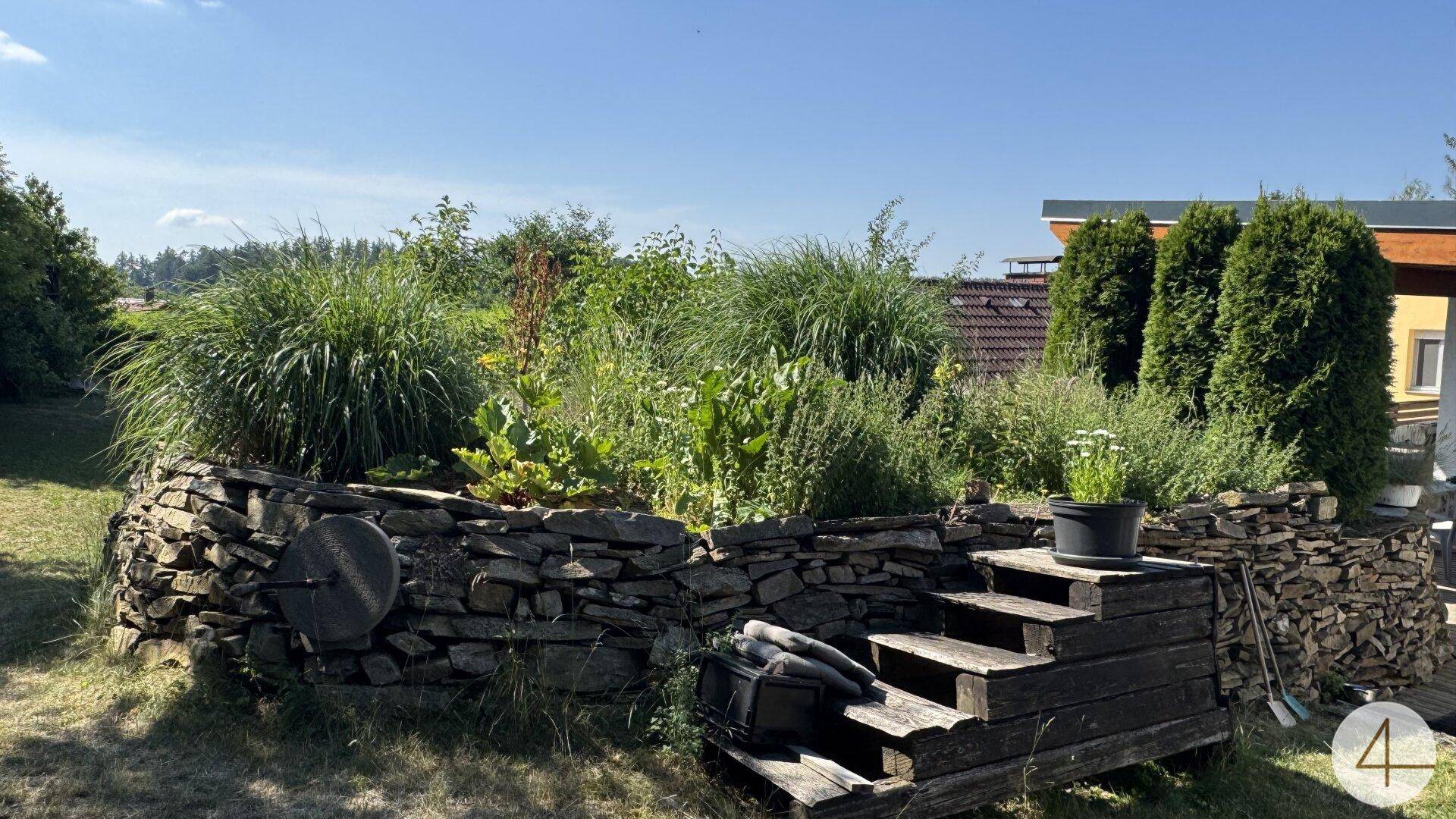 Erhöhtes Kräuter- und Gemüsebeet aus Natursteinen mit Holzstufen in einem sonnigen Gartenbereich.