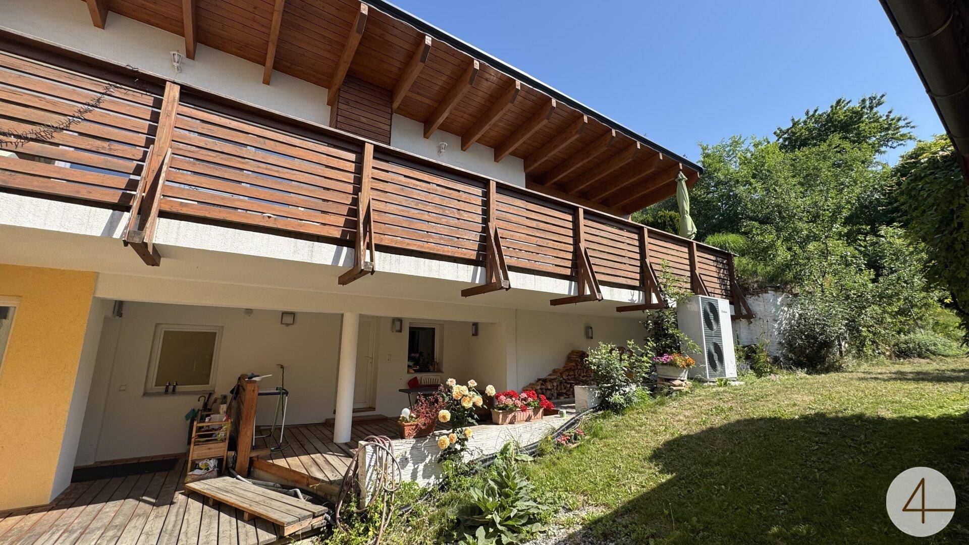 Geräumiger Holzbalkon mit Geländer und darunterliegender Terrasse, umgeben von Pflanzen und Grünflächen.