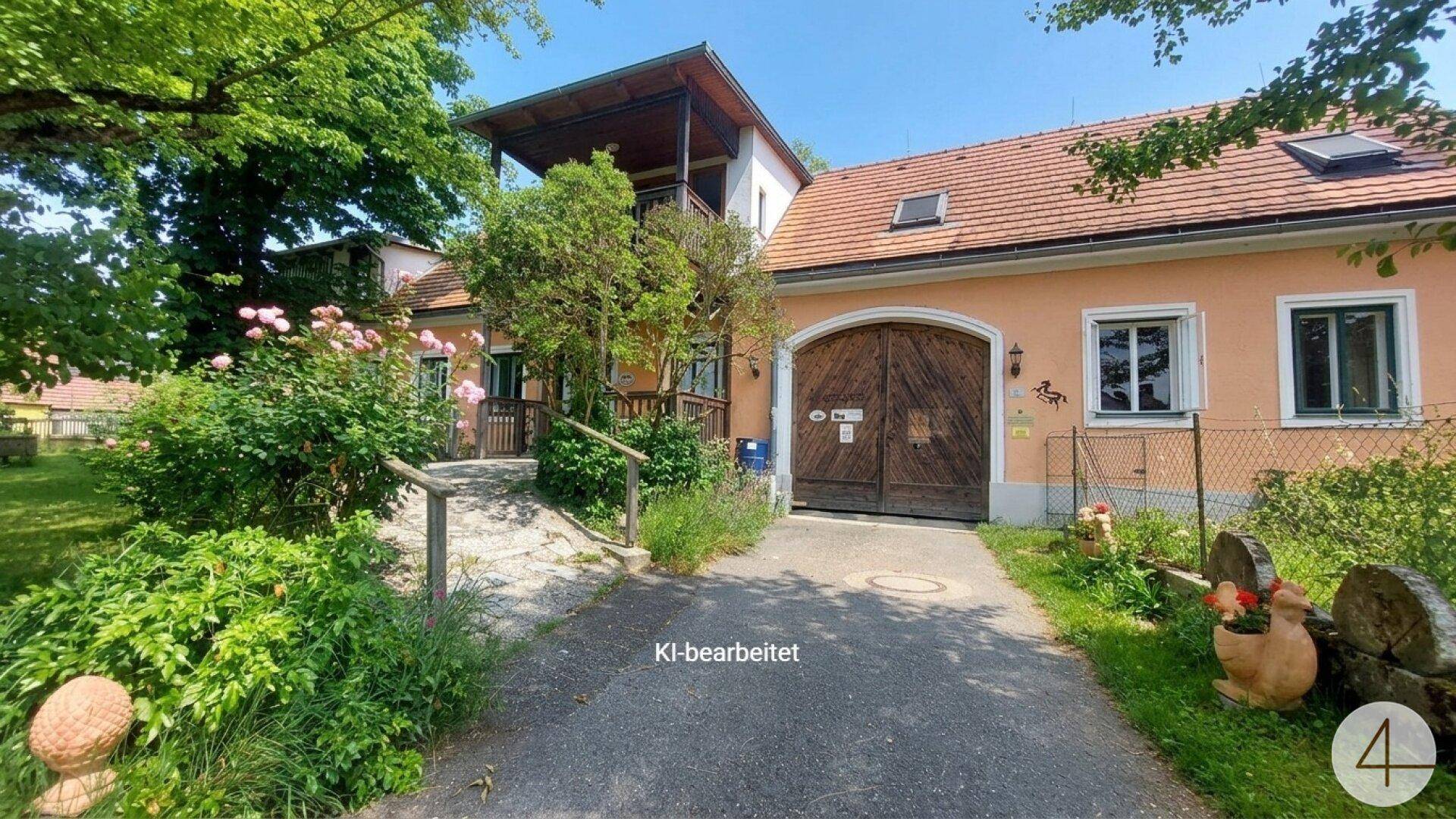 Ansicht des Hauses mit breiter Einfahrt, großem Holztor und einem Balkon im oberen Stockwerk, umgeben von Natur.