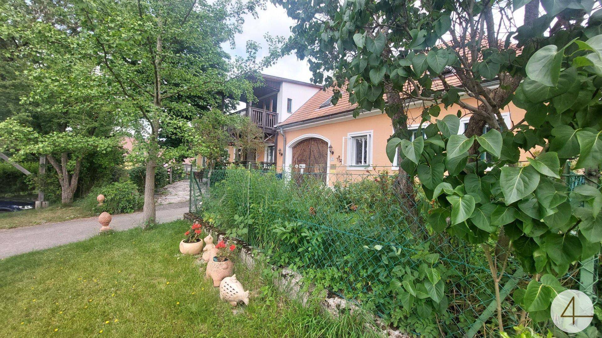 Seitenansicht des Landhauses mit üppigem Garten, hohen Bäumen und Blick auf die Umgebung.