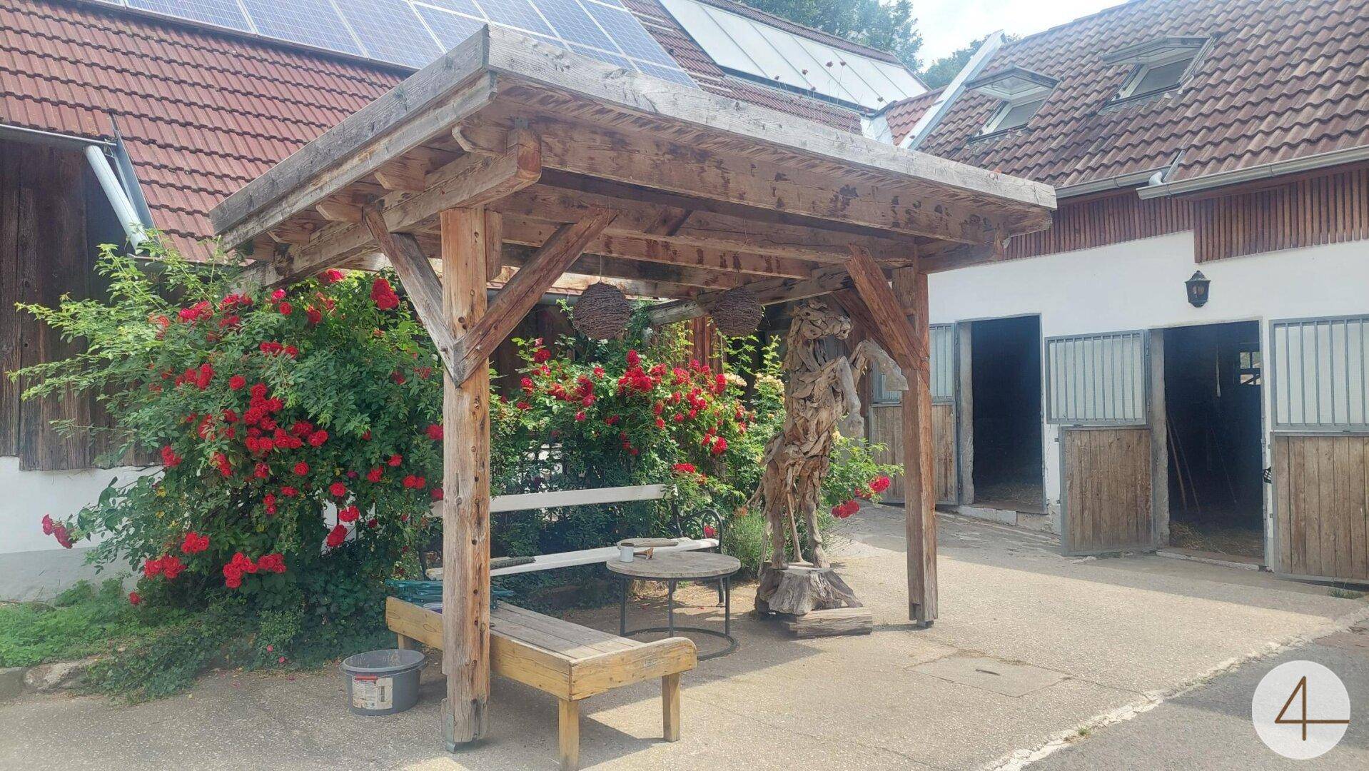 Überdachter Sitzbereich mit Holzkonstruktion, umgeben von roten Rosen, und Solarpaneelen auf dem Dach im Hintergrund.