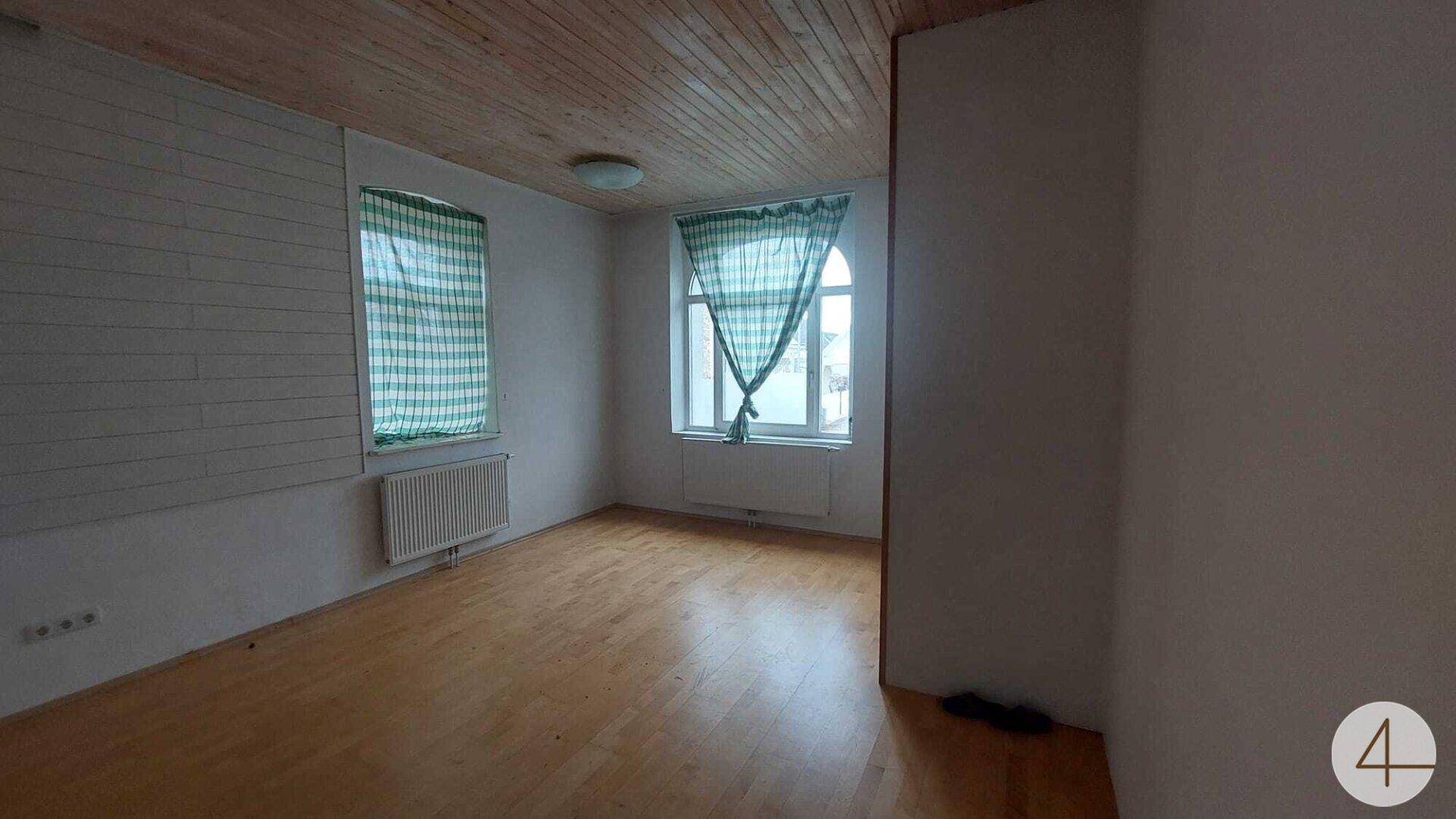 Leerer Raum mit Holzdecke, Holzwandverkleidung und Holzboden, beleuchtet durch zwei Fenster mit karierten Vorhängen.