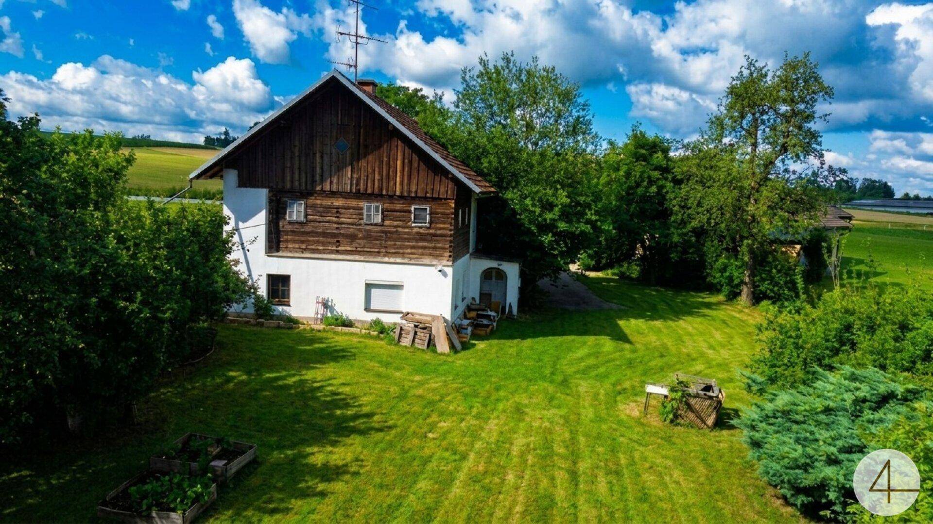Ländliches Haus mit Holzfassade und weißem Sockel, umgeben von einem weitläufigen grünen Garten unter blauem Himmel.