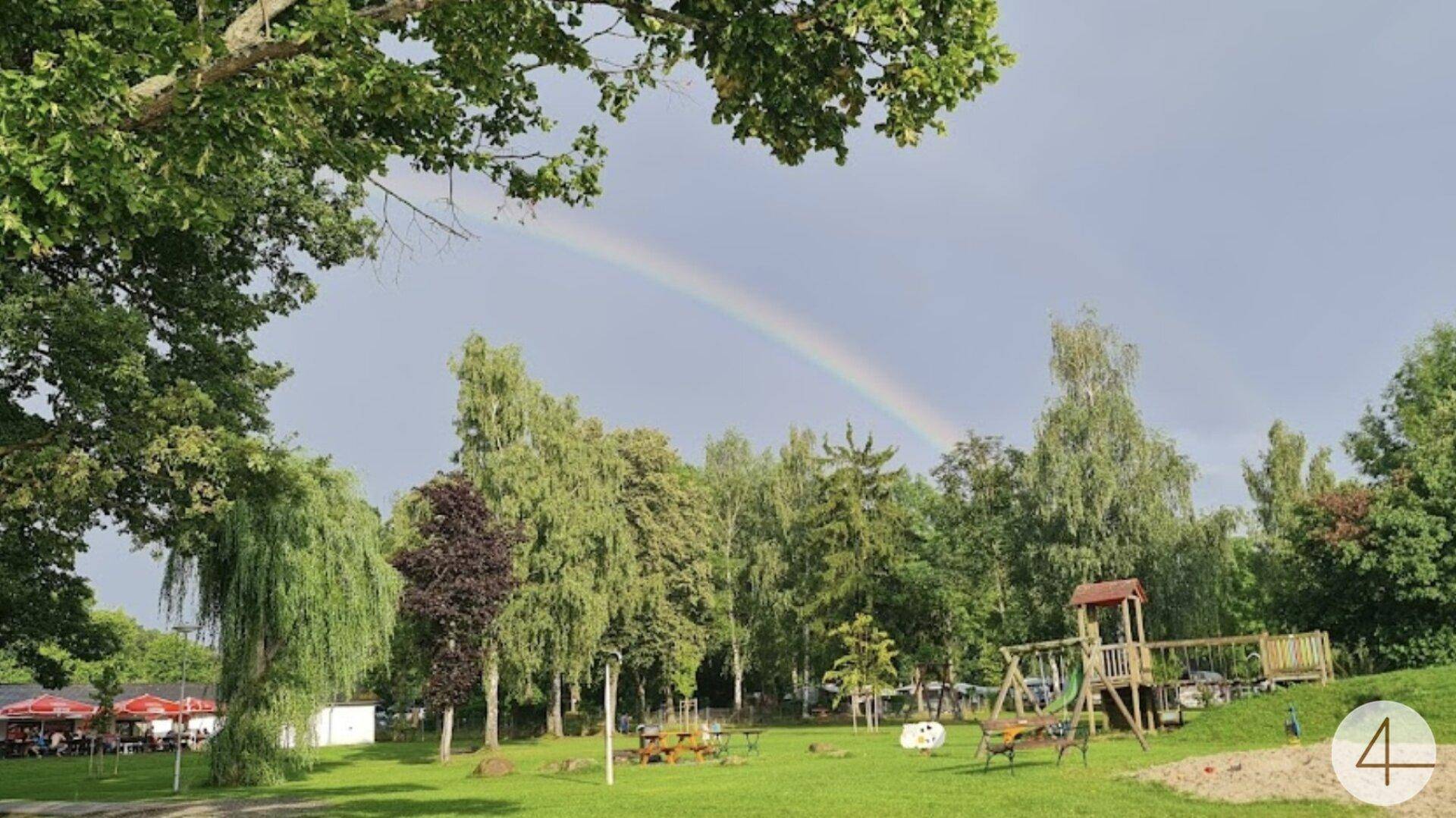 Weitläufiger grüner Garten mit Spielplatz und Bäumen unter einem Regenbogen am Himmel.