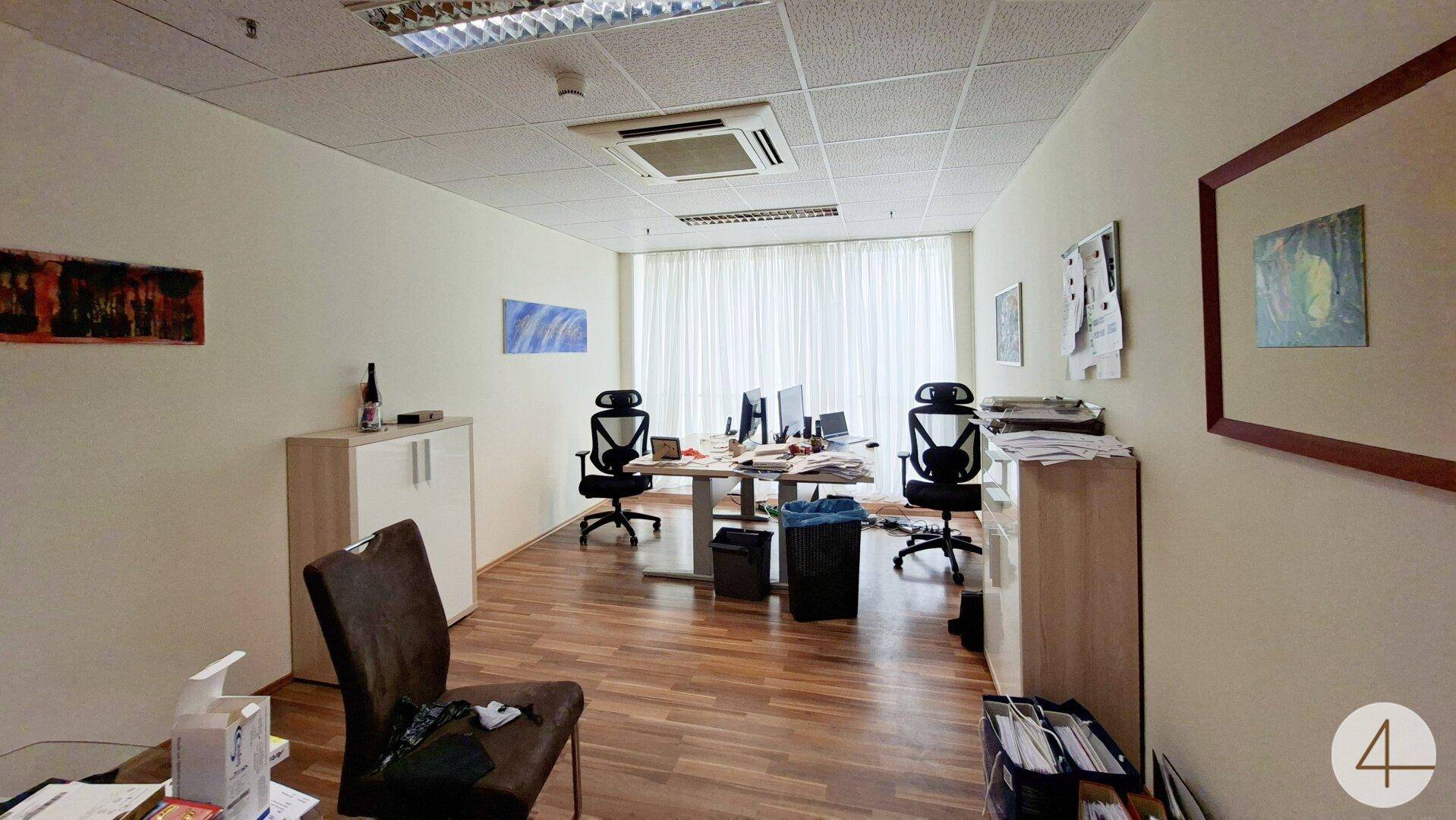 Helles Büro mit mehreren Arbeitsplätzen, Schreibtischen, Bürostühlen und großen Fenstern mit Vorhängen.