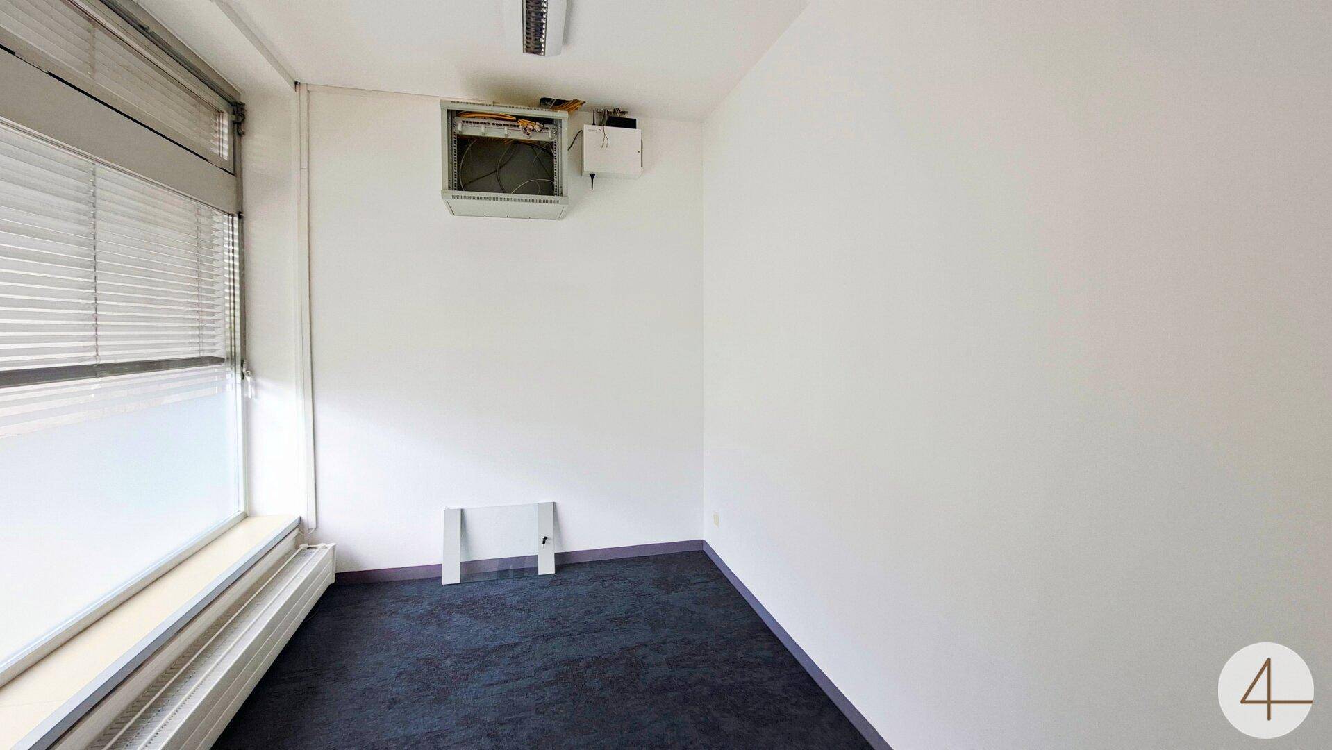 Kleiner Raum mit dunklem Teppichboden, Fenster mit Jalousien und sichtbarer Klimaanlage.