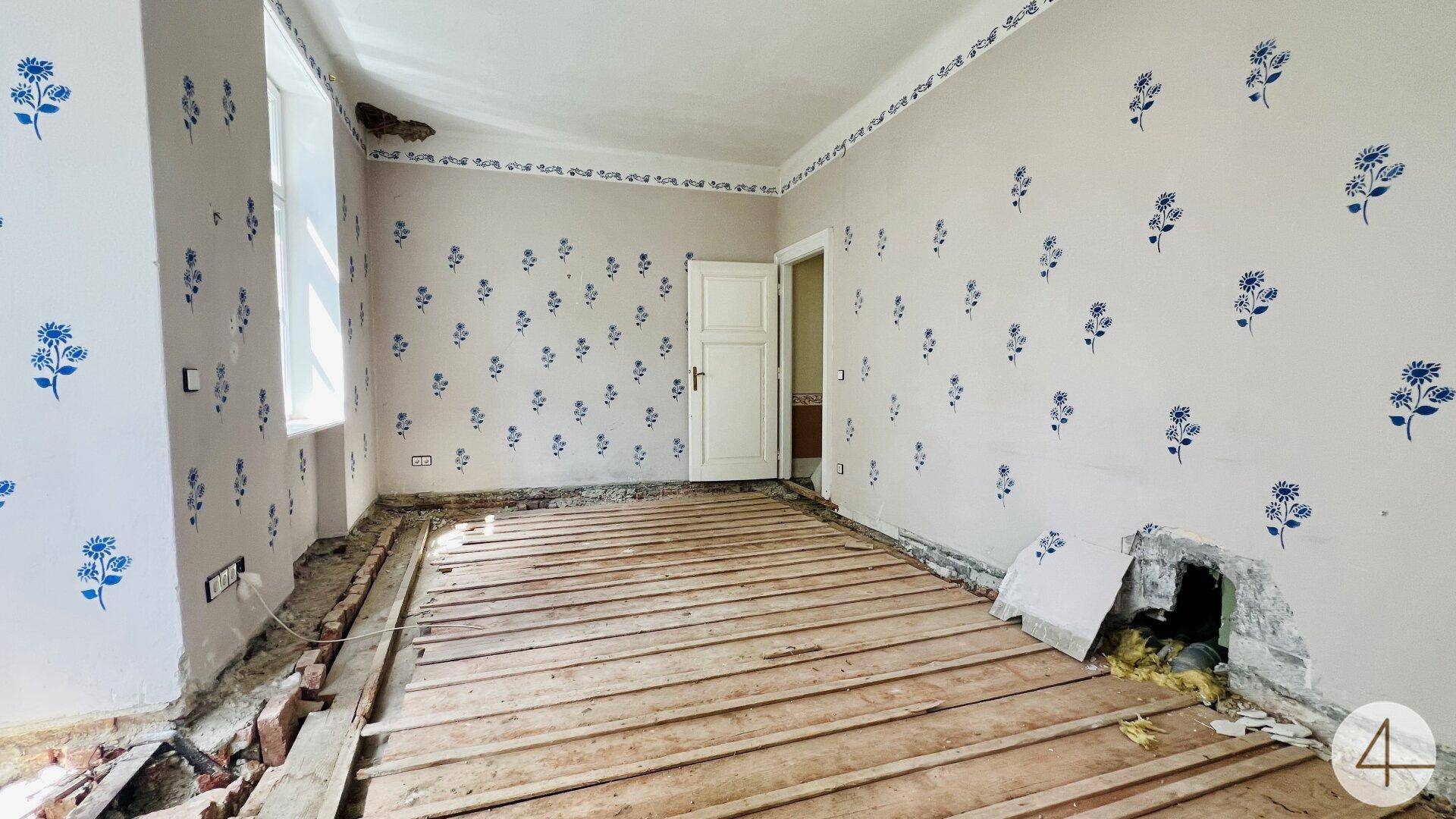 Helles Zimmer mit weiß-blauer Blumenmuster-Tapete und freigelegten Holzdielen, bereit zur Renovierung.