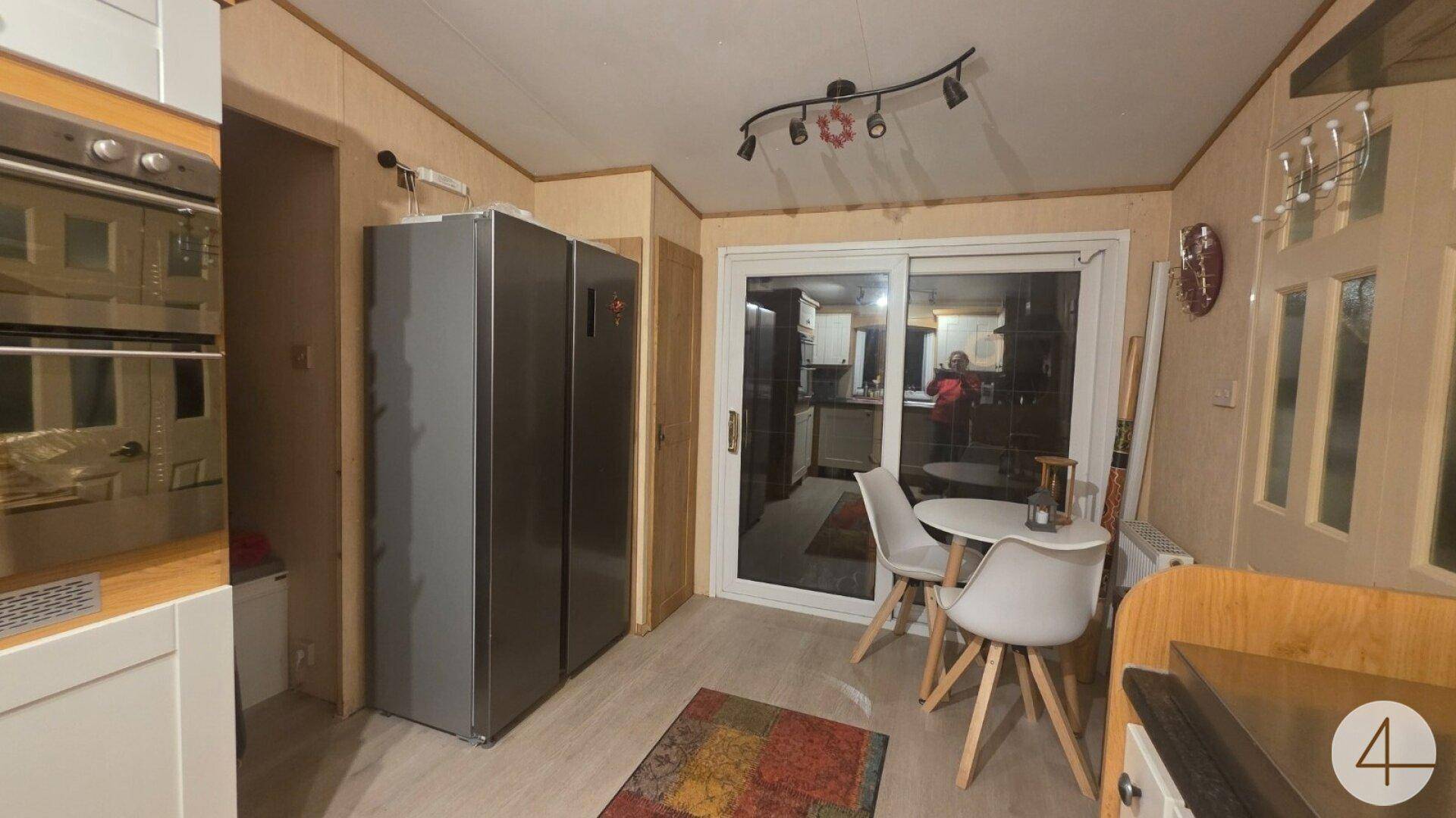 Küchenbereich mit modernen Kühlschränken, Backofen und einem kleinen Esstisch am Fenster.