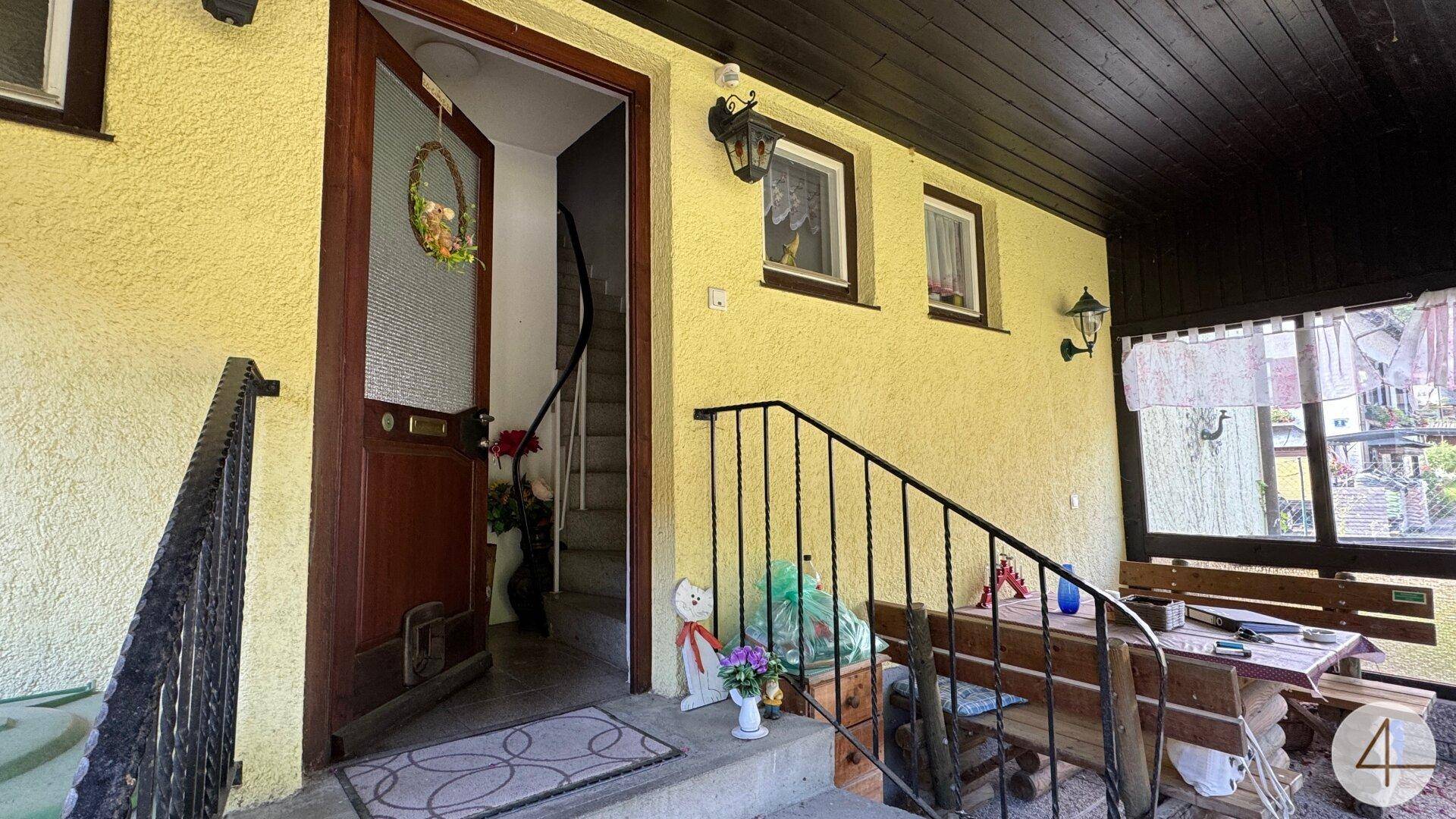 Überdachter Eingangsbereich des Hauses mit offener Tür, die den Blick auf eine Innentreppe freigibt.