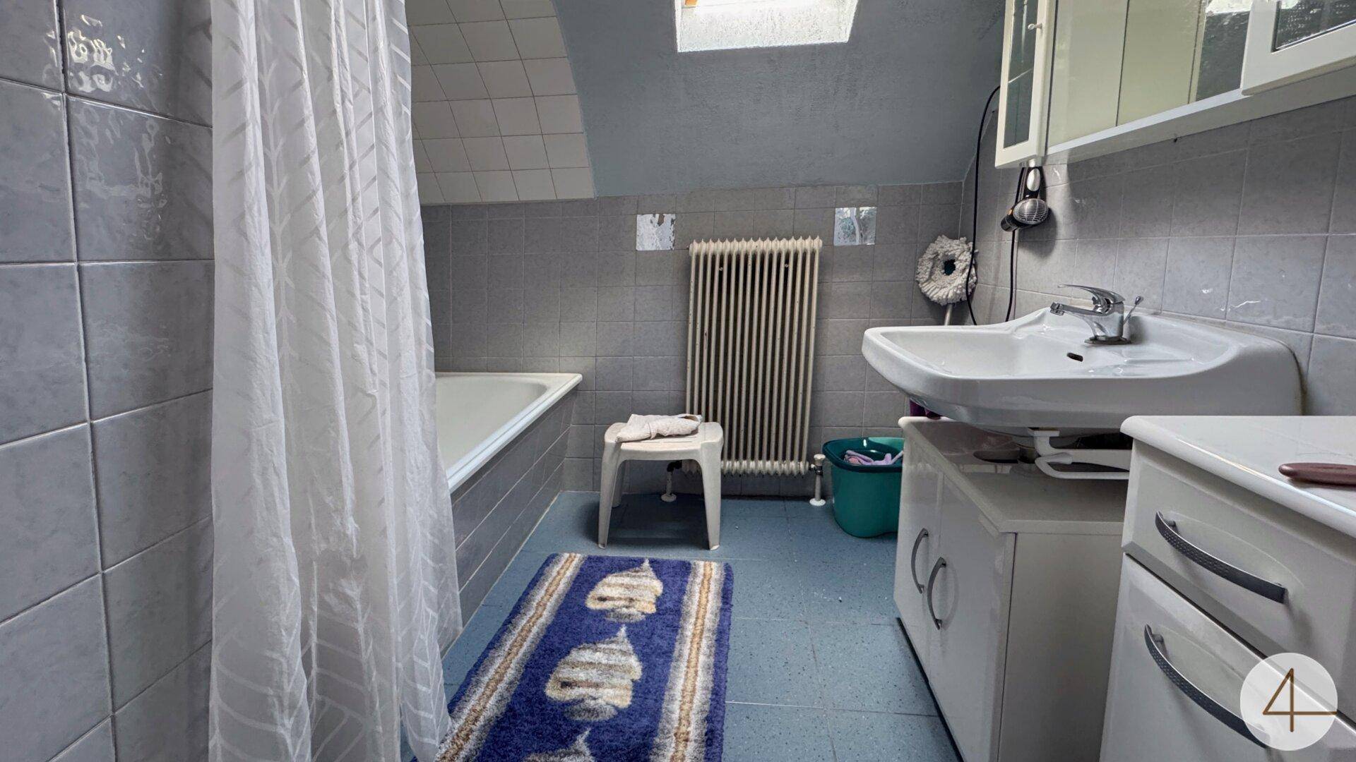 Badezimmer mit grauen Fliesen, Badewanne mit Duschvorhang, Waschbecken und einem Dachfenster.