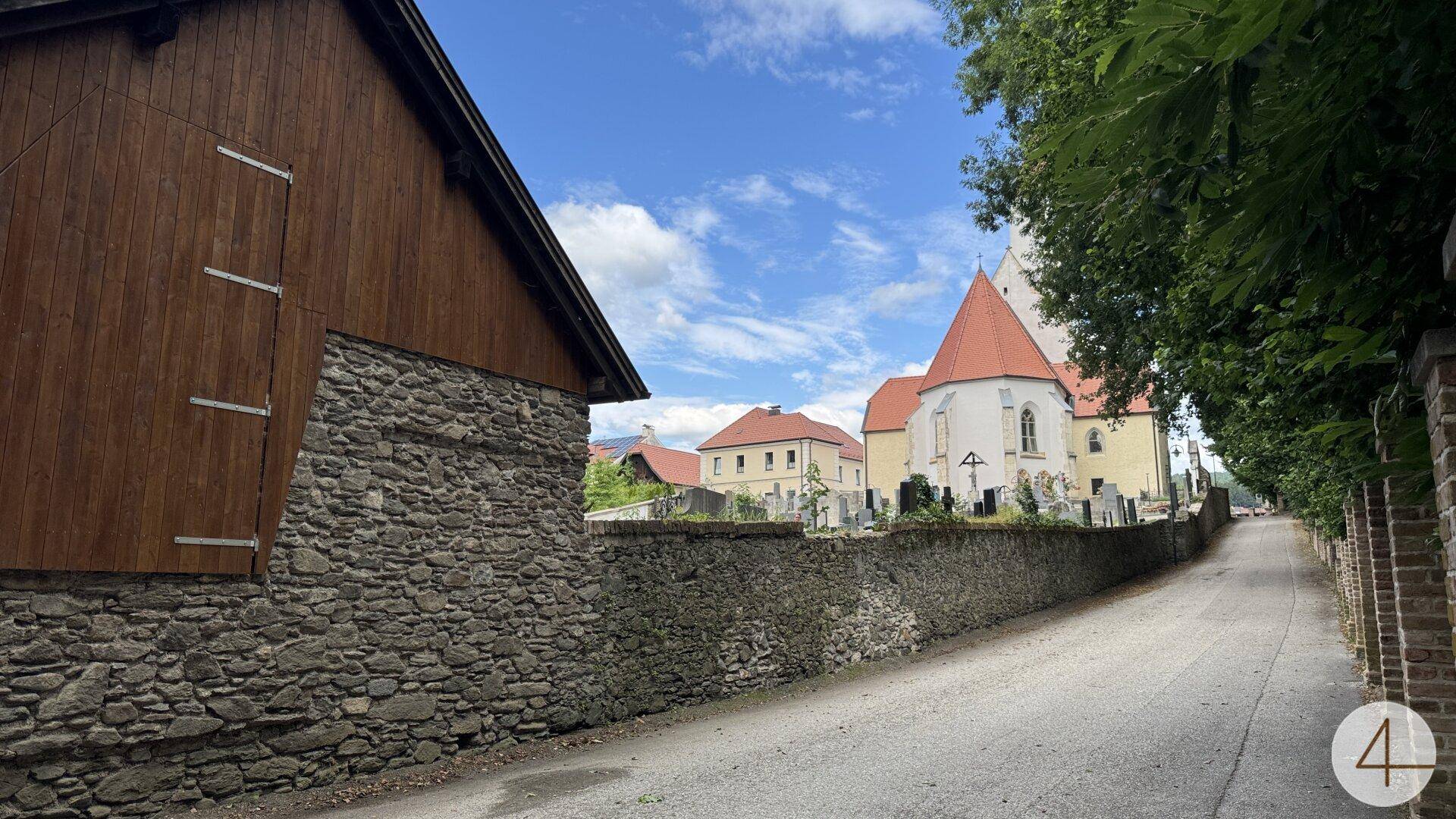 Historische Dorfstraße mit alten Steinmauern, einer Scheune und einer Kirche im Hintergrund.