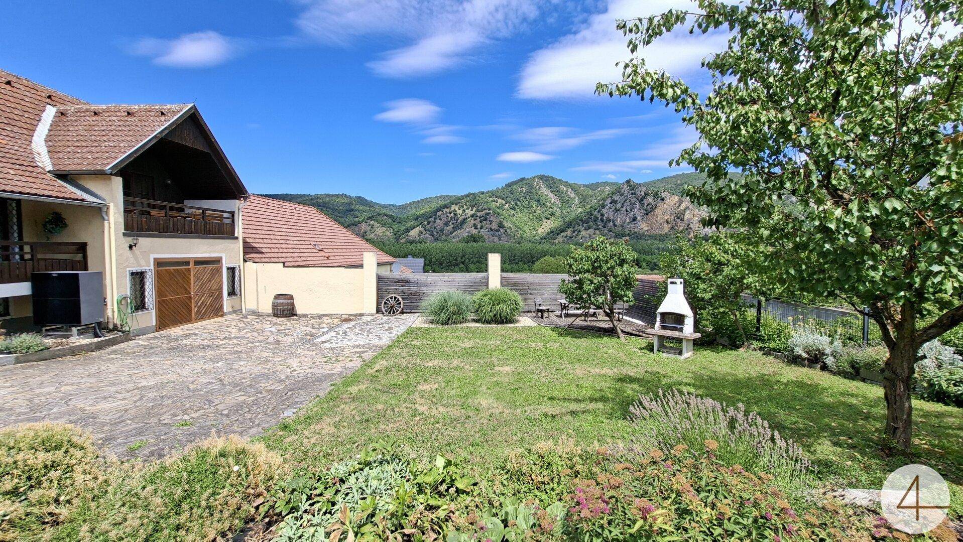 Sonniger Garten mit Grillplatz und Sitzgelegenheiten, umgeben von grüner Landschaft.