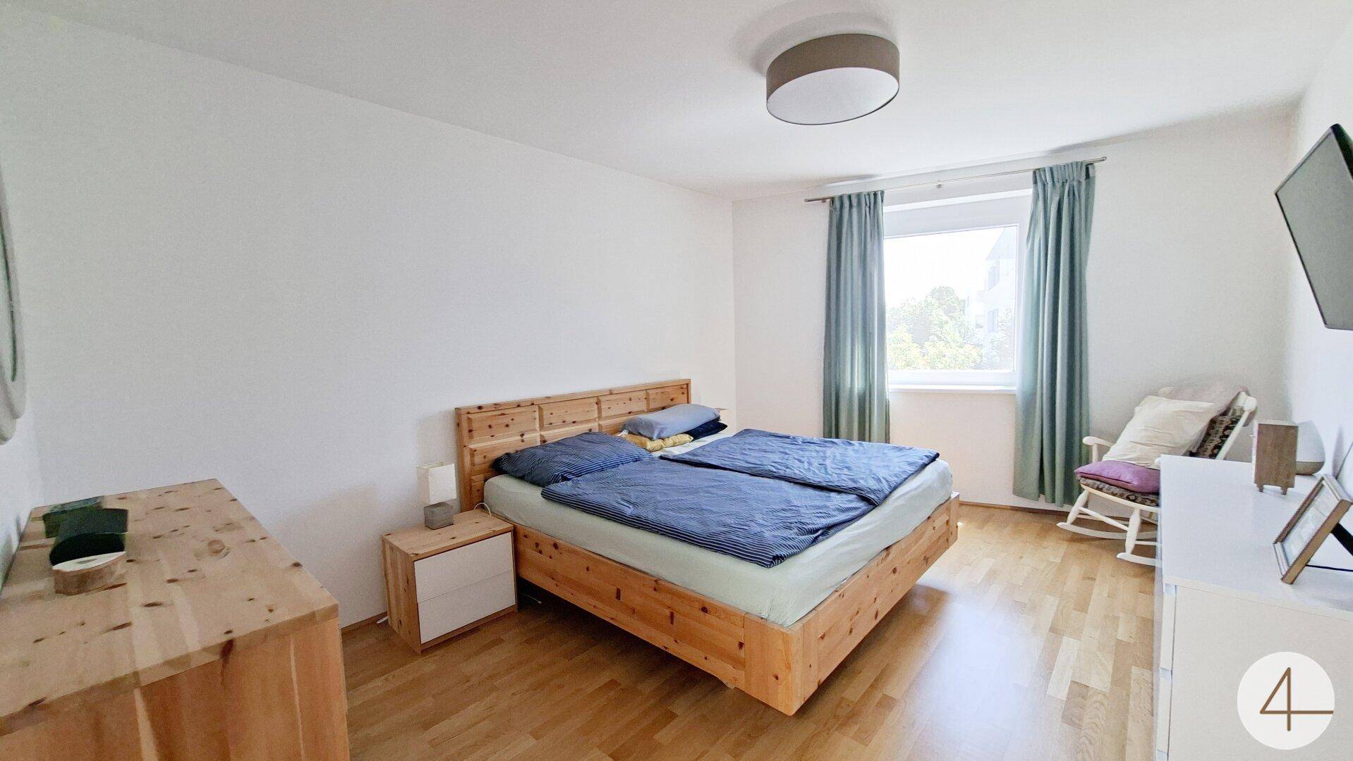 Gemütliches Schlafzimmer mit Holzbett, Holzboden und Fenster mit Vorhängen.