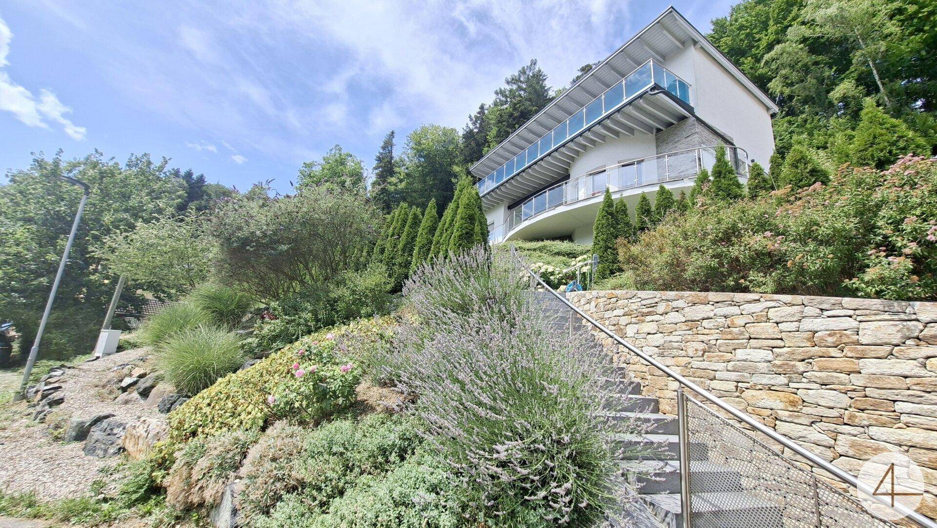 Elegantes Haus in Hanglage mit Steintreppe und üppiger Bepflanzung, umgeben von Natur.