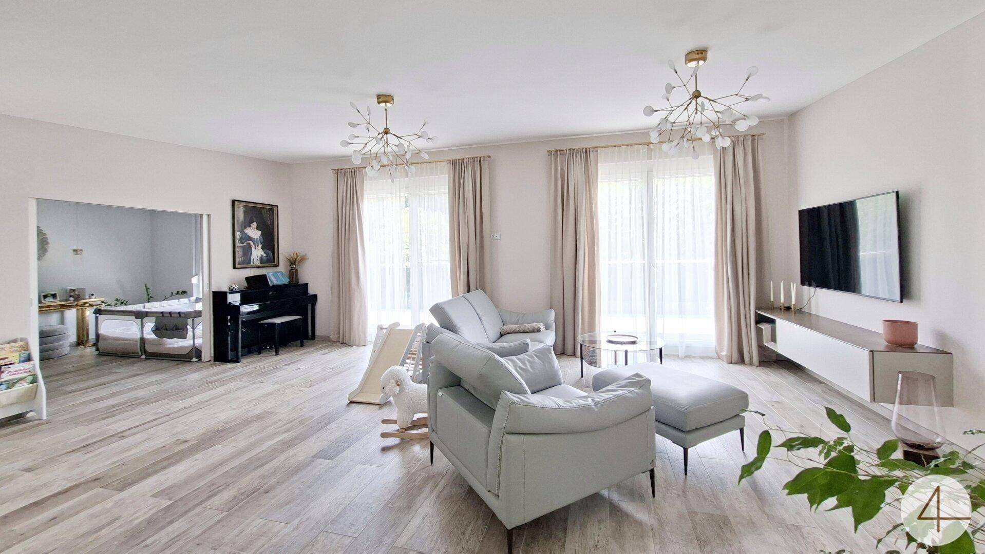 Geräumiges Wohnzimmer mit großen Fenstern, hellen Möbeln und einem Fernseher.