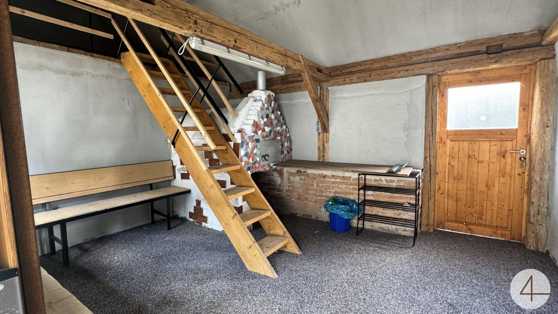 Rustikaler Raum mit Holzbalken, Treppe und gemauertem Ofen, der auf Fertigstellung wartet.