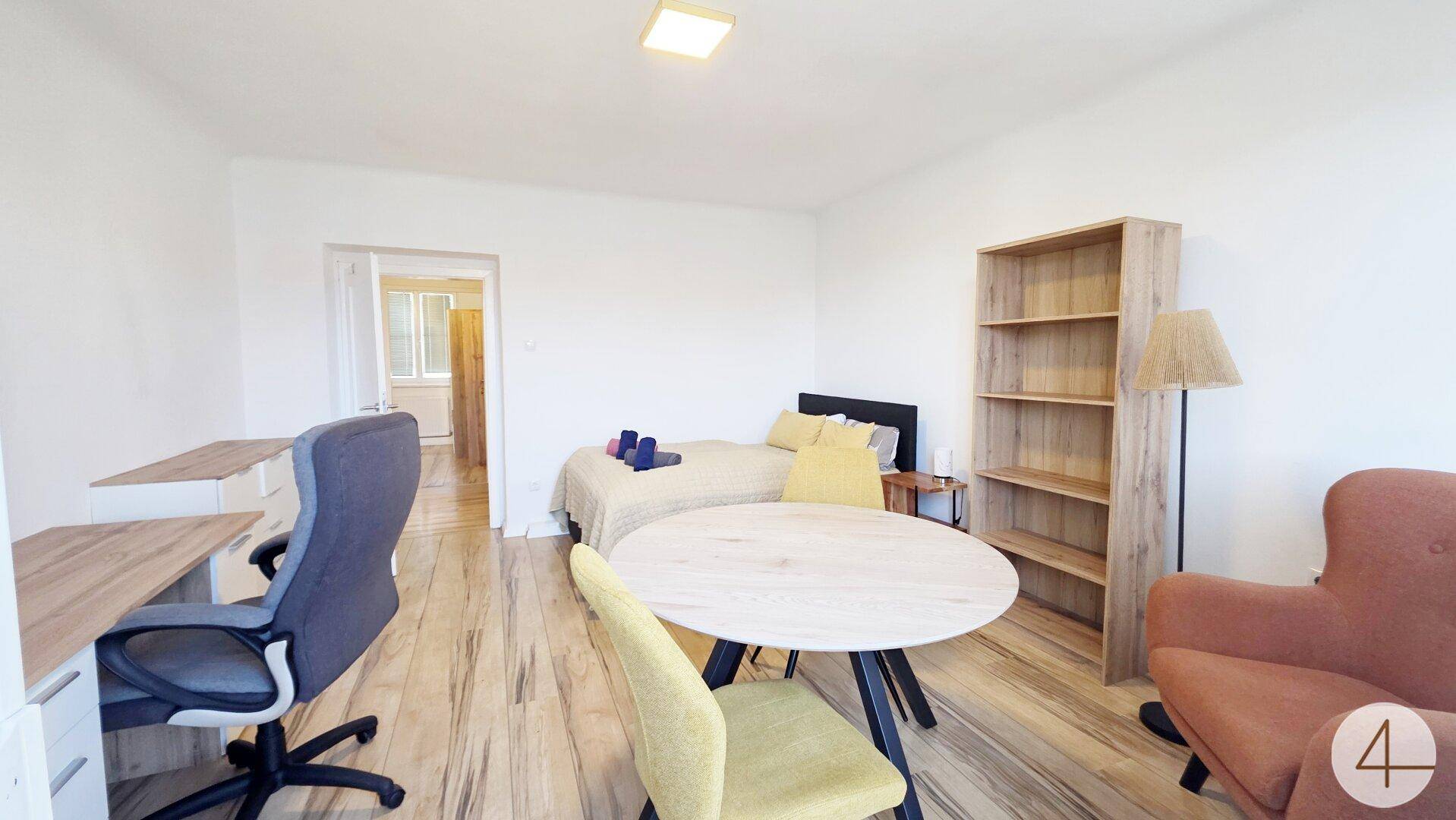 Vielseitiges Schlafzimmer mit Bett, Esstisch, Schreibtisch und Blick in einen weiteren Raum.