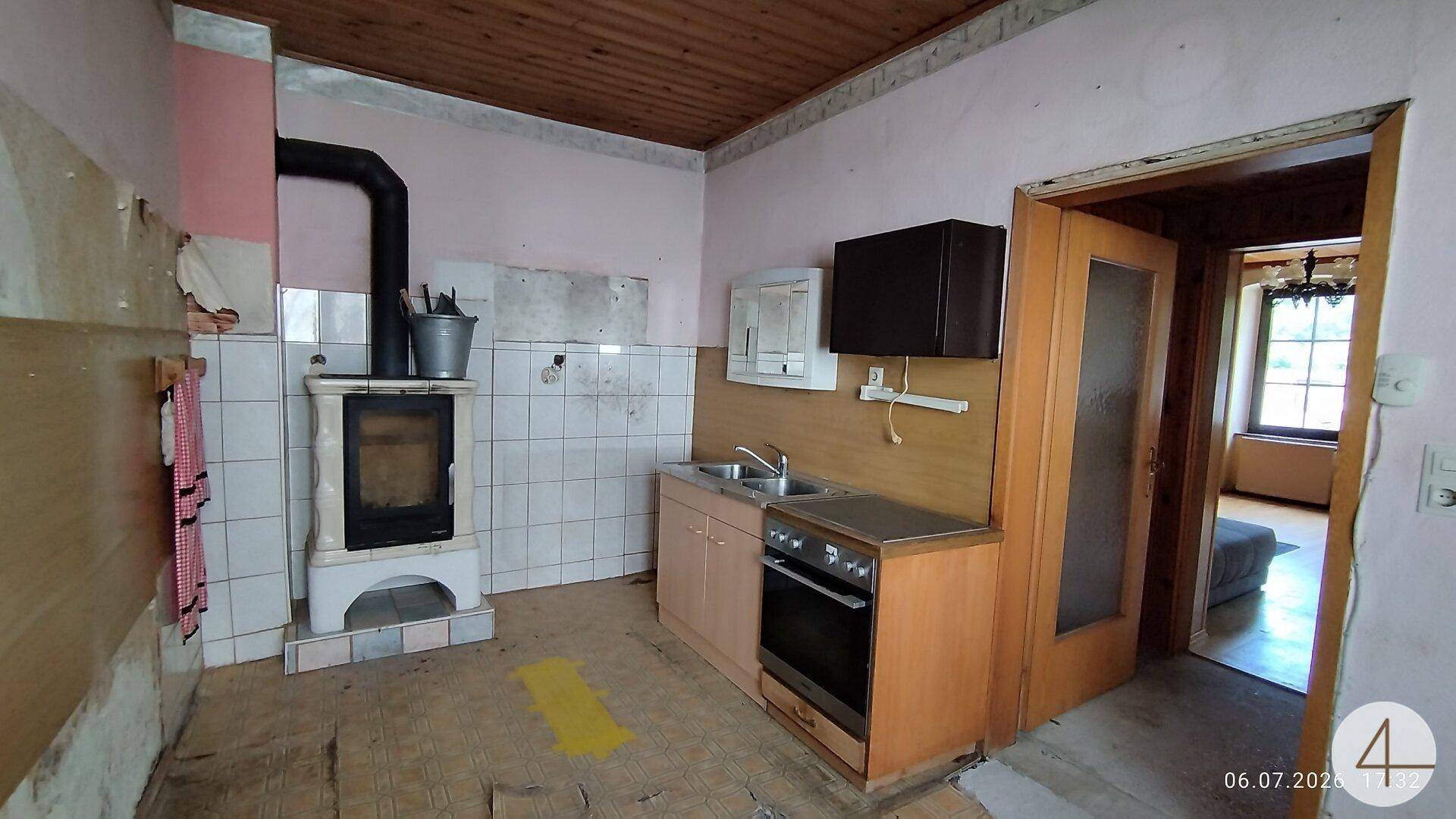 Renovierungsbedürftige Küche mit altem Holzofen, einfacher Spüle und Blick in einen weiteren Raum.