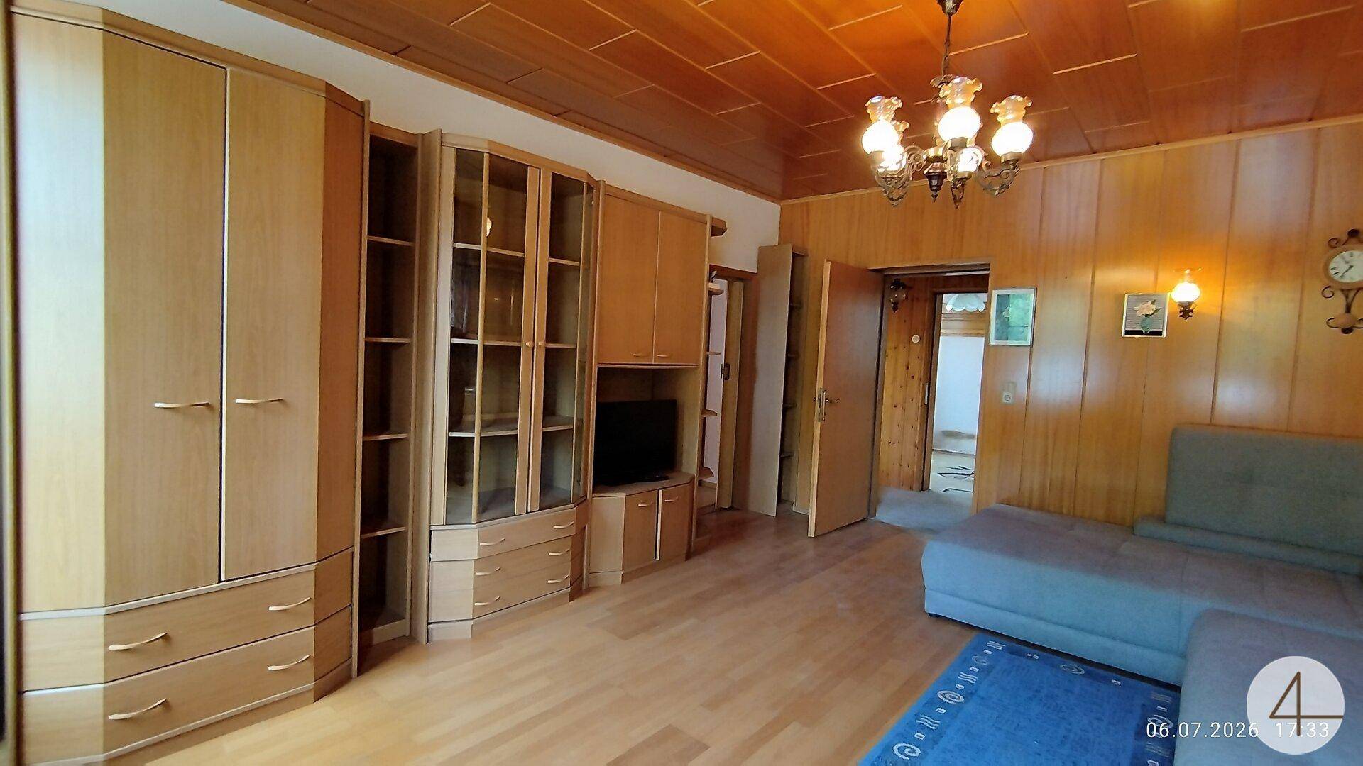 Wohnzimmer mit Holztäfelung, Schrankwand und Sofa, das zu einem weiteren Raum führt.