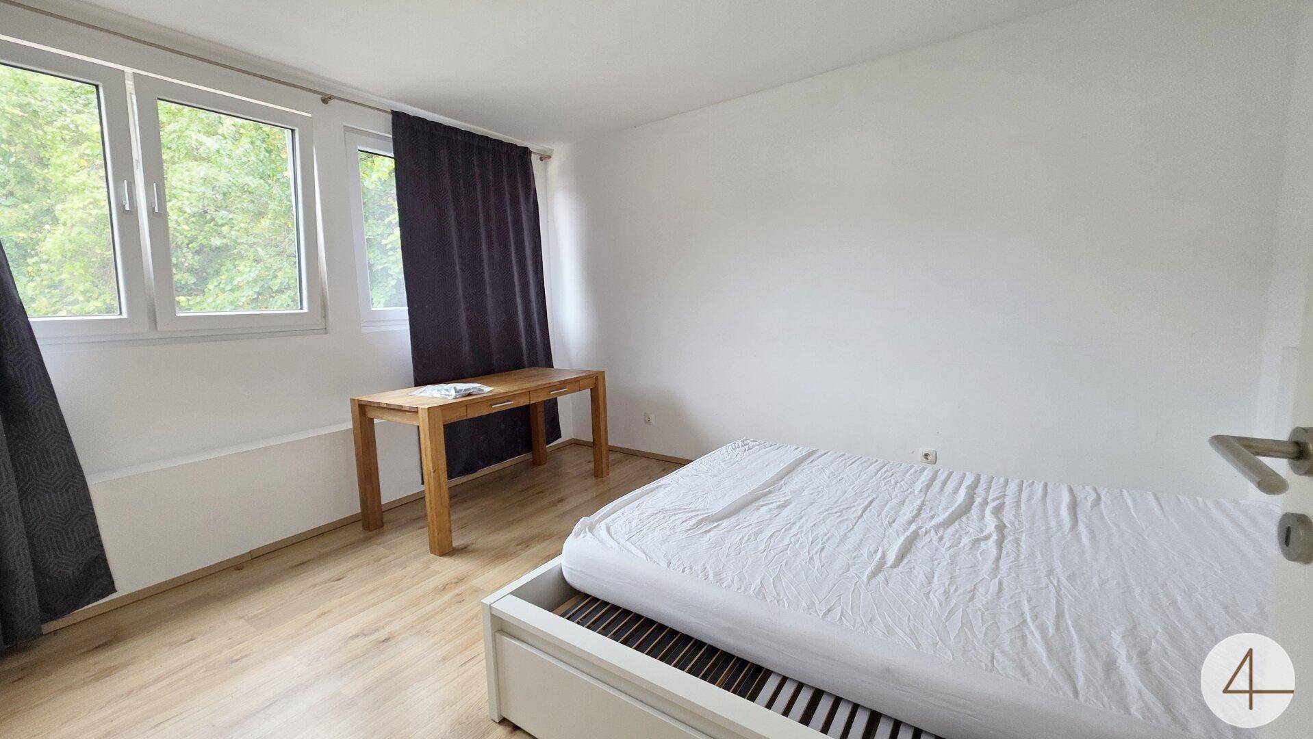 Helles Schlafzimmer mit Holzboden, einem Bett und einem Schreibtisch unter großen Fenstern.