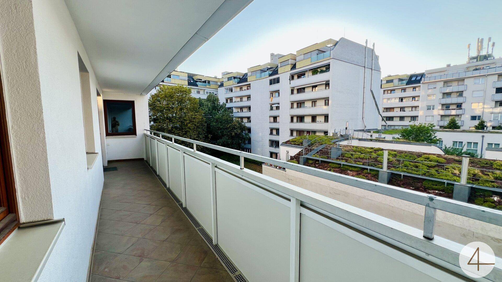 Heller Balkon mit Fliesen und Blick auf die Stadt sowie grüne Dächer der Nachbargebäude.