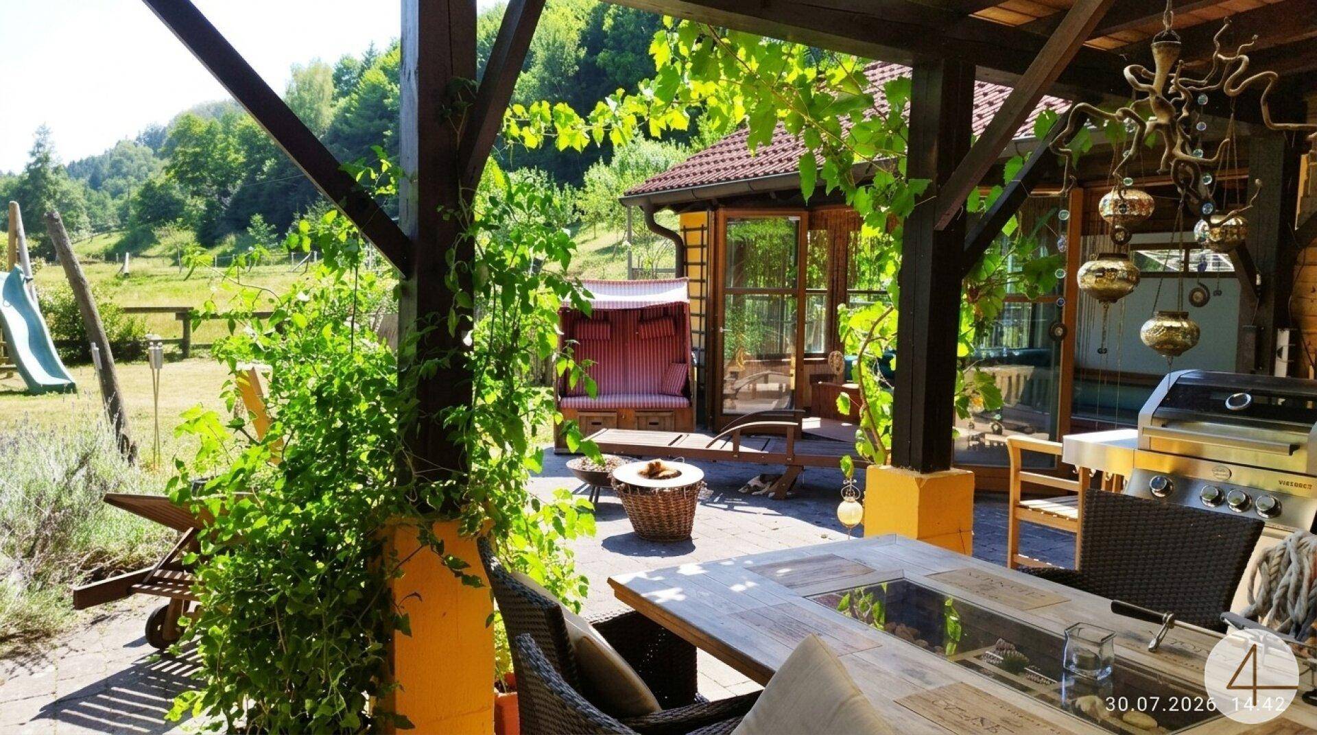 Überdachte Terrasse mit Esstisch, Grillbereich und Blick auf den Garten mit Spielplatz und grüner Landschaft.