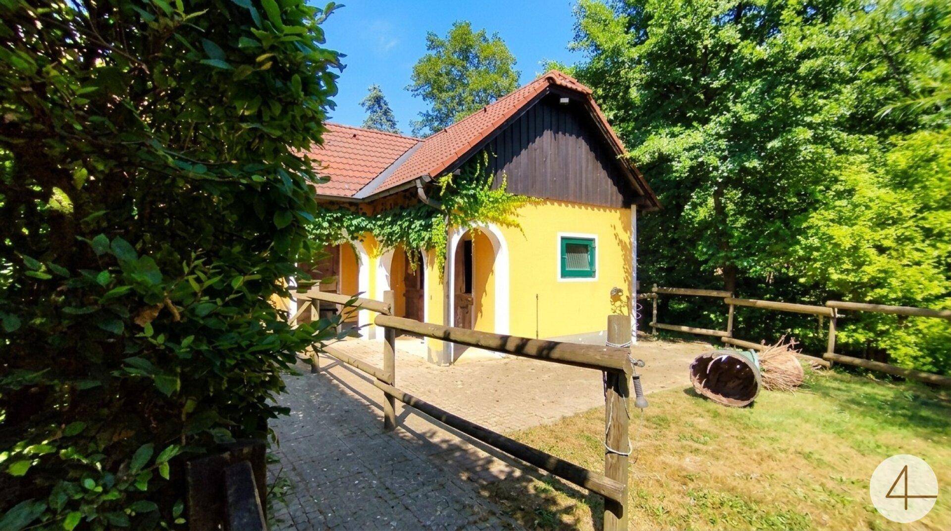 Charmantes gelbes Nebengebäude mit rotem Ziegeldach, umgeben von üppigem Grün und Holzzaun.